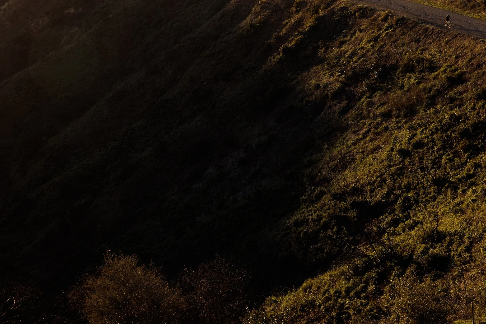 A winding mountain road with a cyclist riding on it, surrounded by green hills and vegetation, illuminated by sunlight.