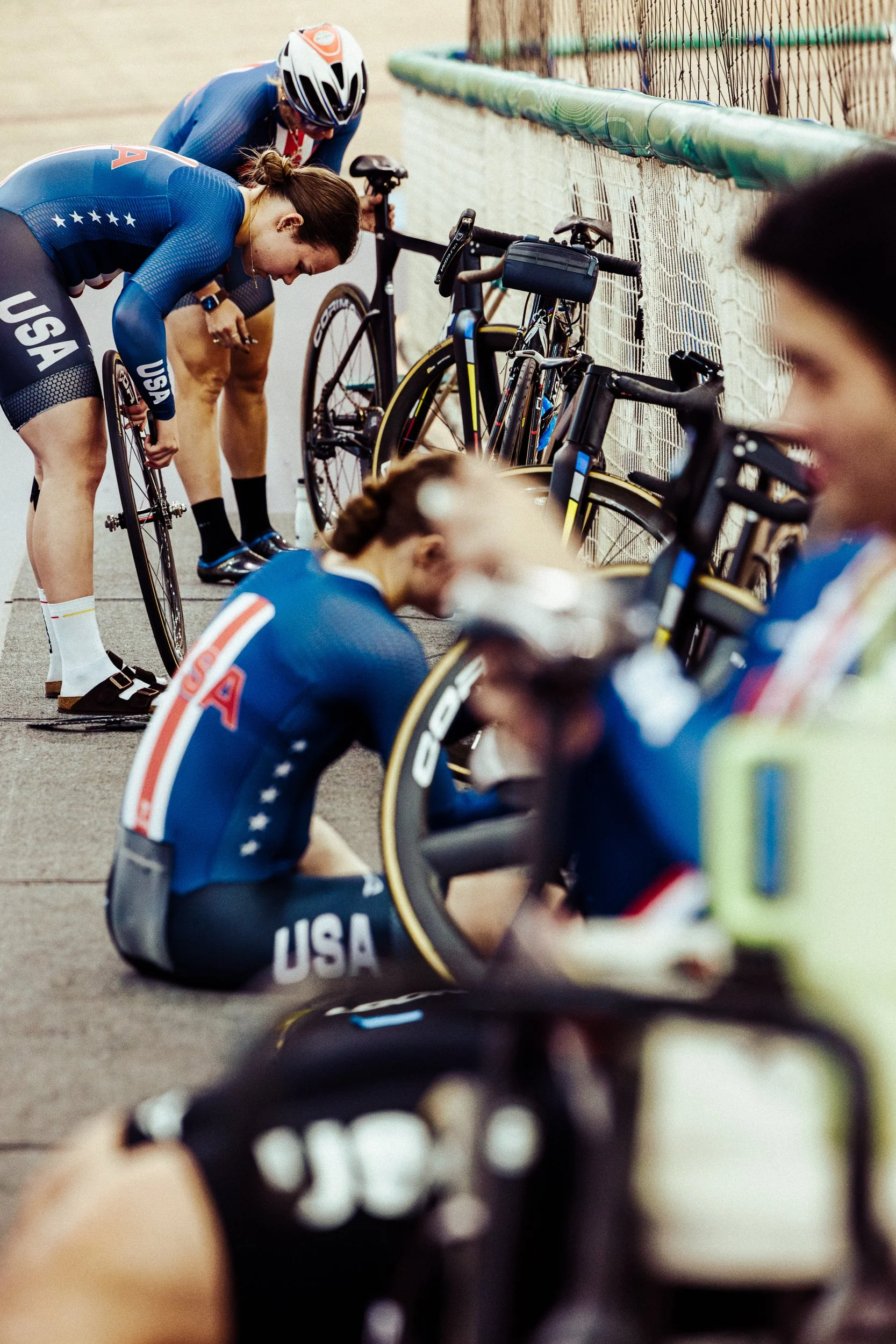 U.S. female track cyclists preparing for a race, bent over their bikes, with additional team members in the background, inside a velodrome.
