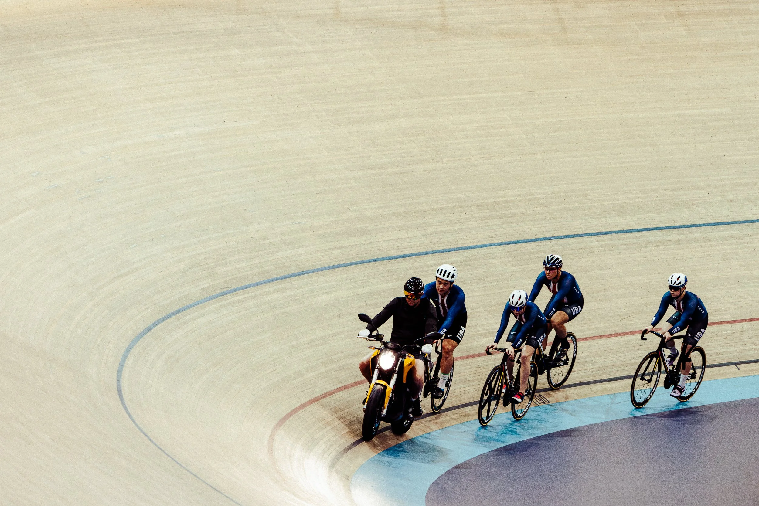 A coach on a motorcycle leading four cyclists racing inside a velodrome.