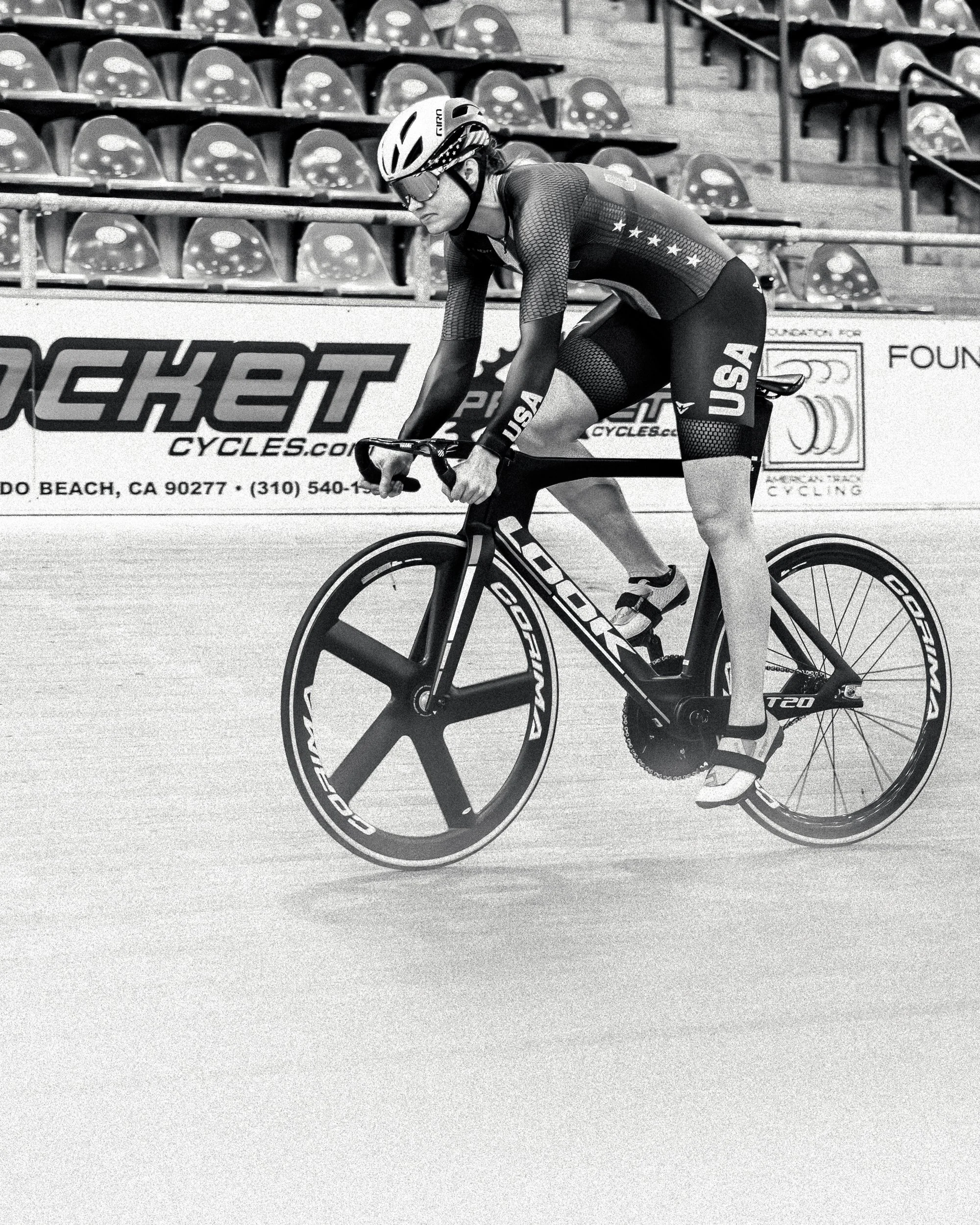 Black and white photo of a female cyclist in racing gear and helmet riding a time trial bike on a velodrome track with empty bleachers in the background.