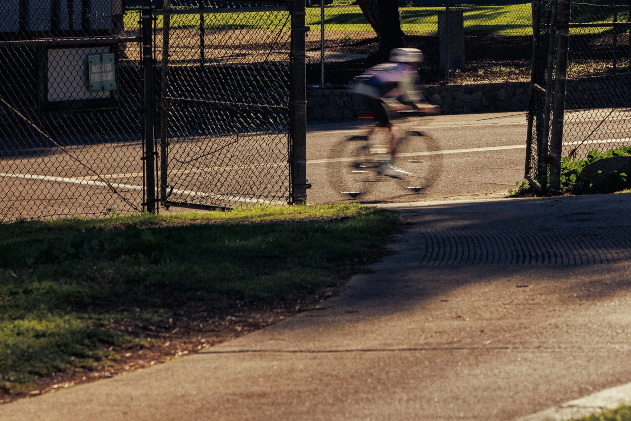 A person riding a bicycle through a park gate with a sign that says "Watch for pedestrians". The person is blurred due to motion, and the scene is illuminated by sunlight.