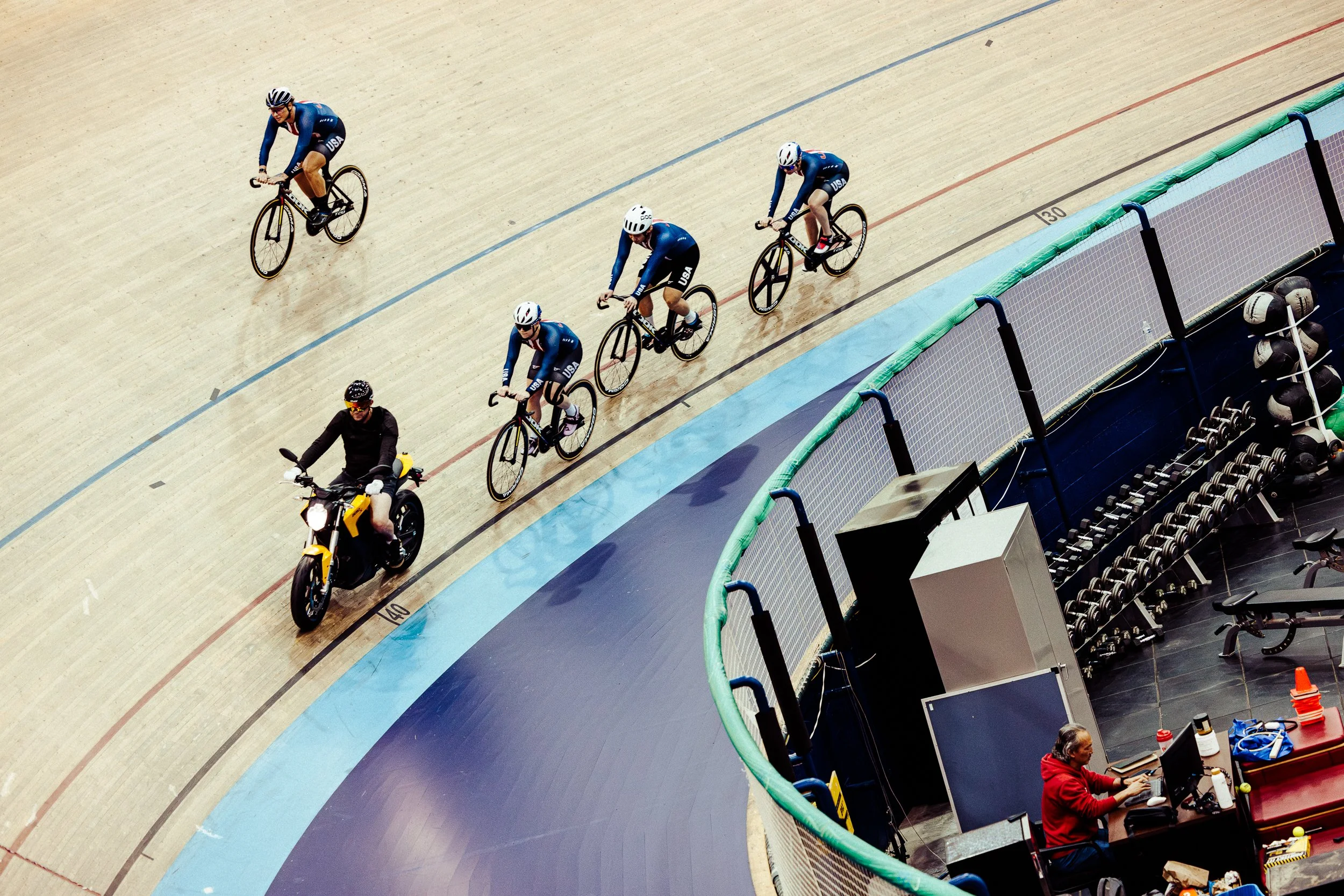 Cyclists competing in a track race, with a motorcycle escort, at an indoor velodrome.