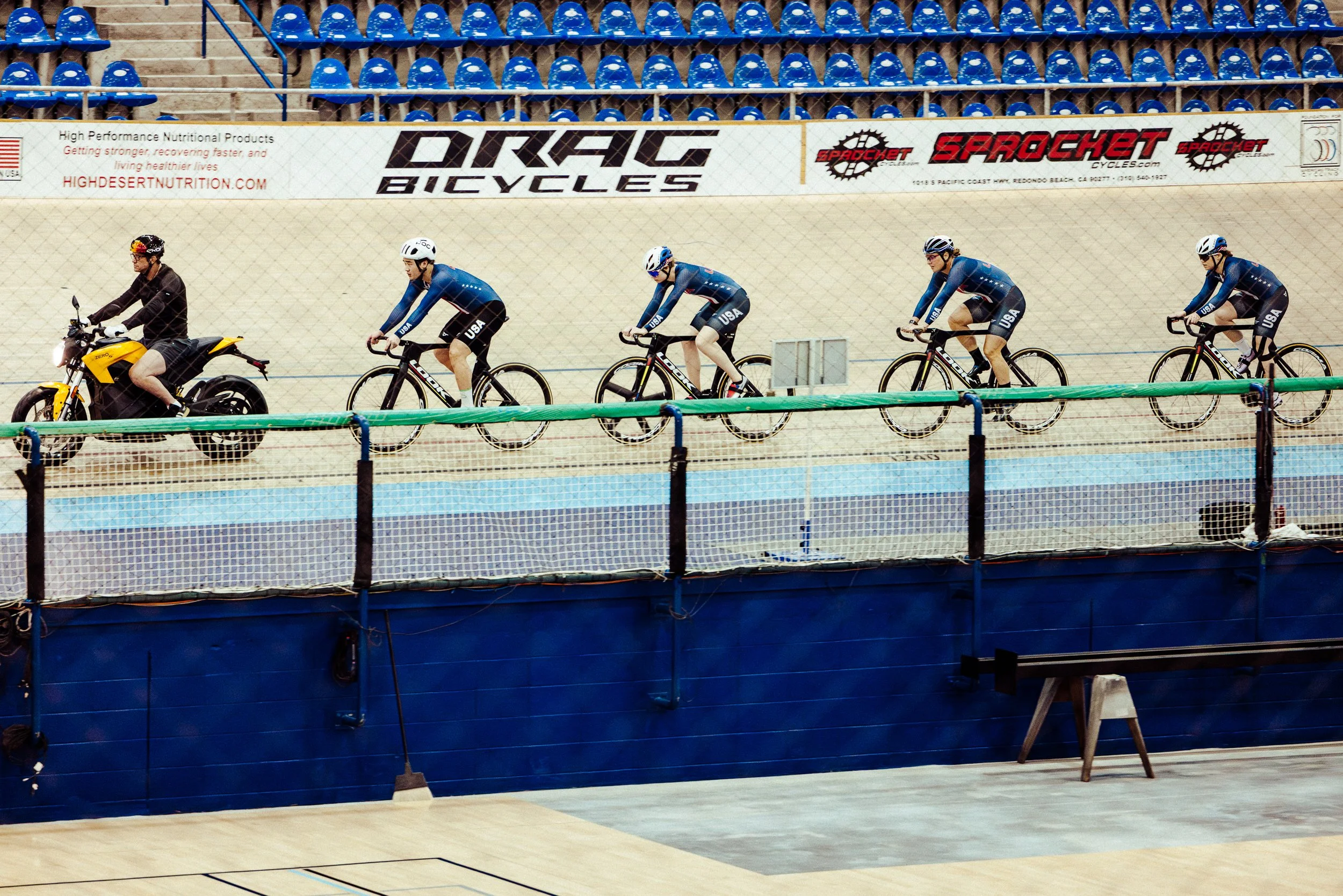 A group of five cyclists in blue uniforms riding track bikes on an indoor velodrome track, led by a person on a yellow motorcycle. The velodrome has blue seats and advertising banners in the background.