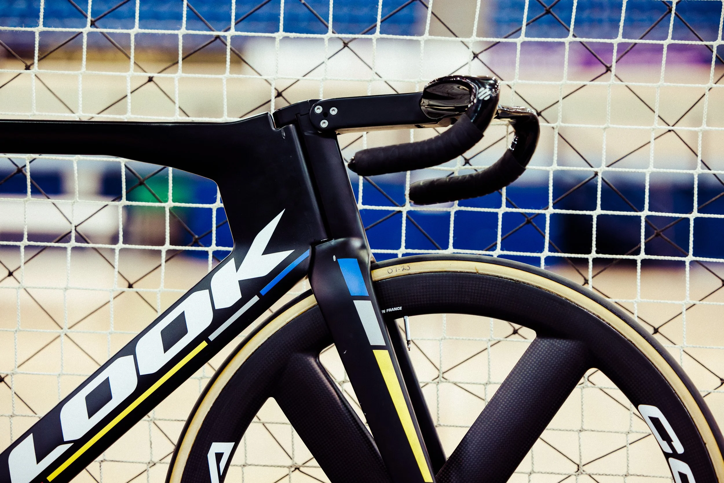 Close-up of a black and yellow racing bicycle with a sleek aerodynamic frame, drop handlebars, and deep-section carbon wheels, positioned in front of a wire fence.