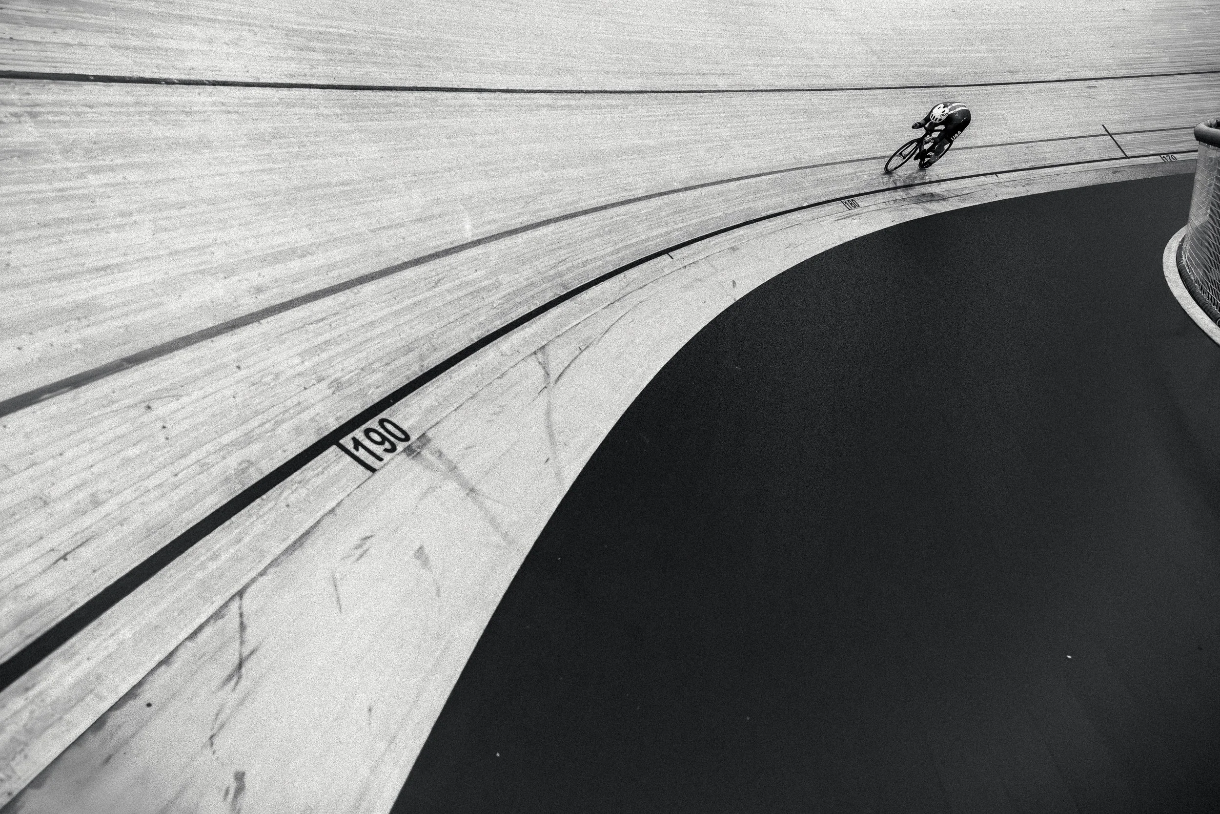 A cyclist riding on a velodrome track, which has markings, including a speed limit of 190. The track has a curved wooden surface with a dark lower section, and the cyclist is wearing a helmet and athletic gear.