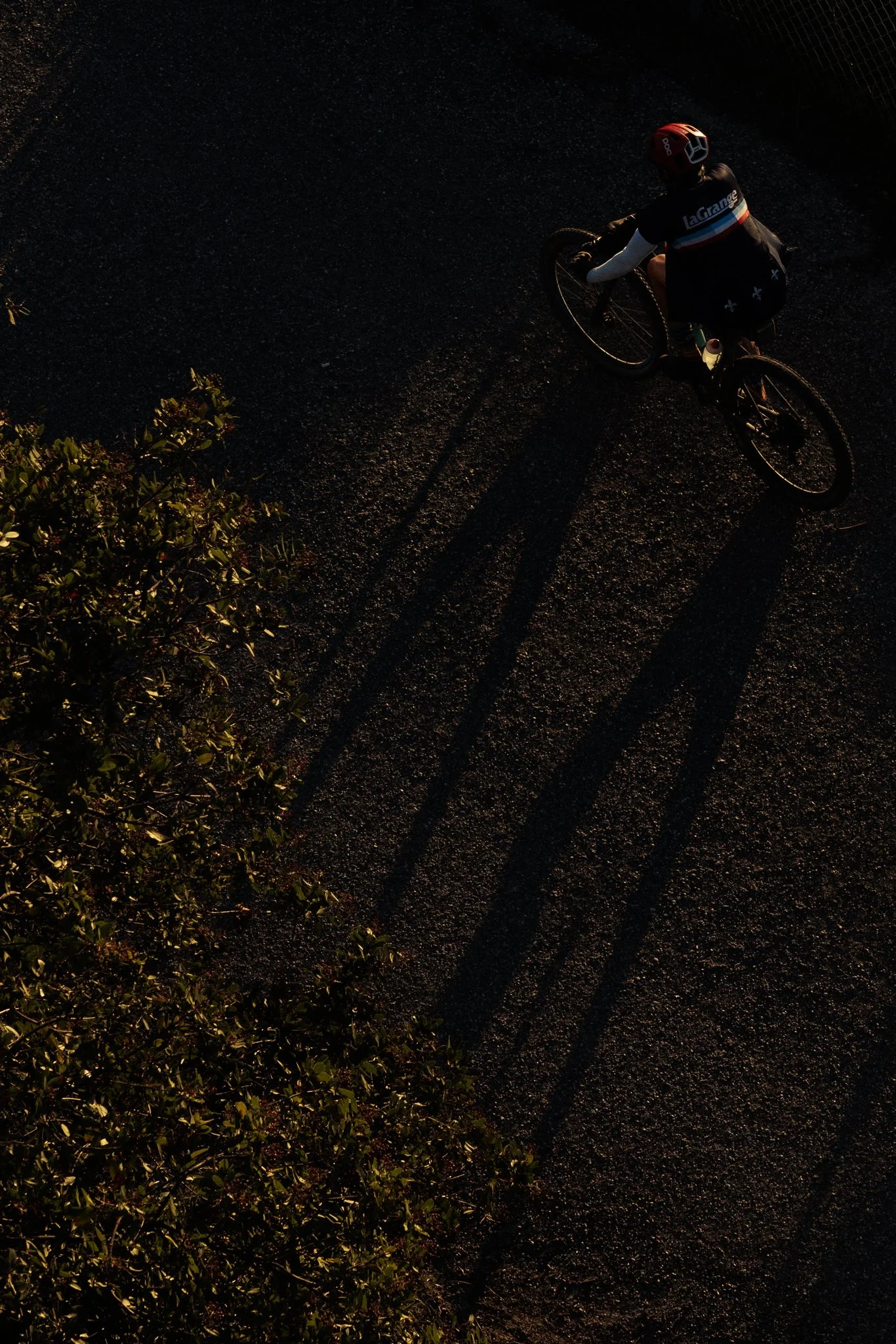 A person riding a bicycle on a dark, gravelly road during sunset, casting long shadows.