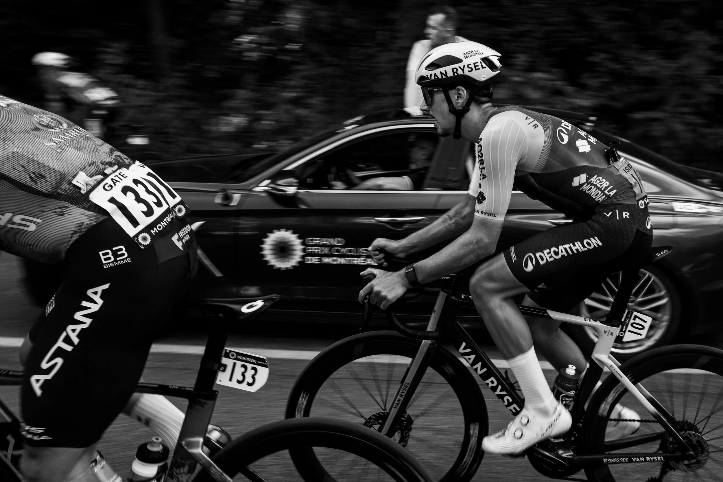 A cyclist in racing gear riding a bike during a race, with a car passing closely behind on a road.