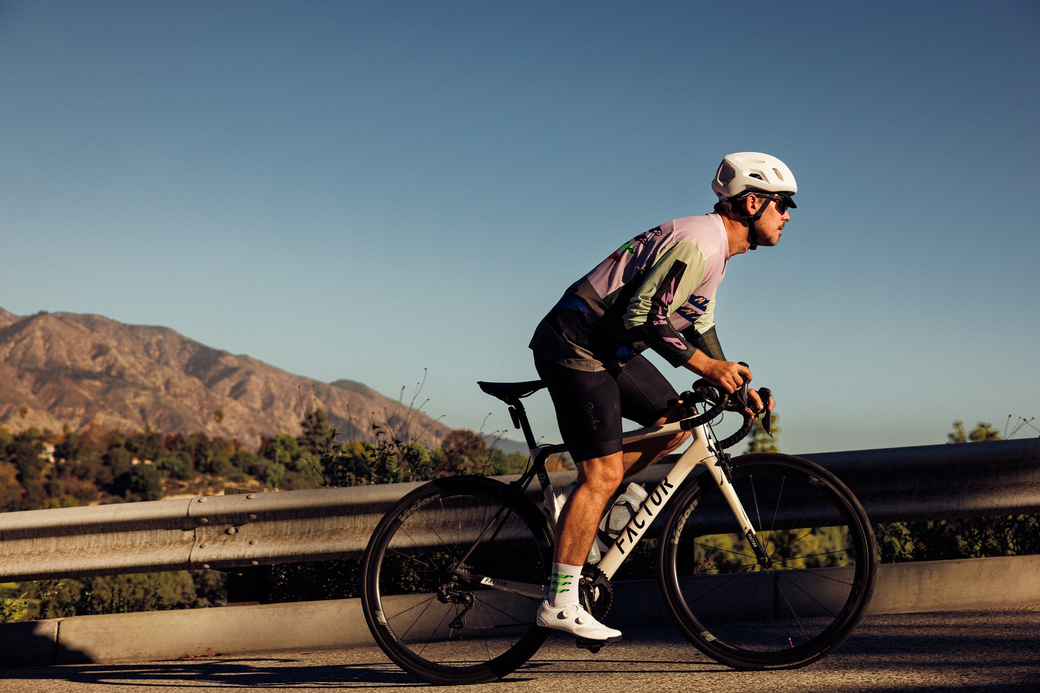 Man riding a white bicycle on a road with mountains in the background, wearing a helmet, sunglasses, a colorful cycling jersey, black shorts, and white shoes during sunset.
