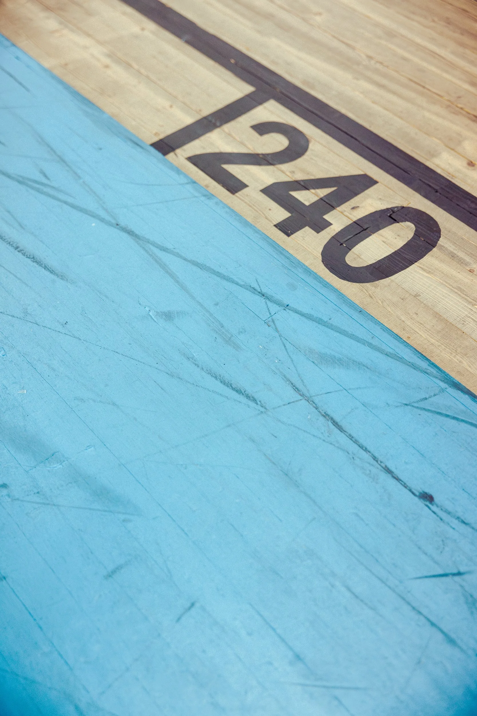 Close-up of a wooden basketball court floor with markings, including the number 240 and black lines. A blue section of the court is visible in the foreground.