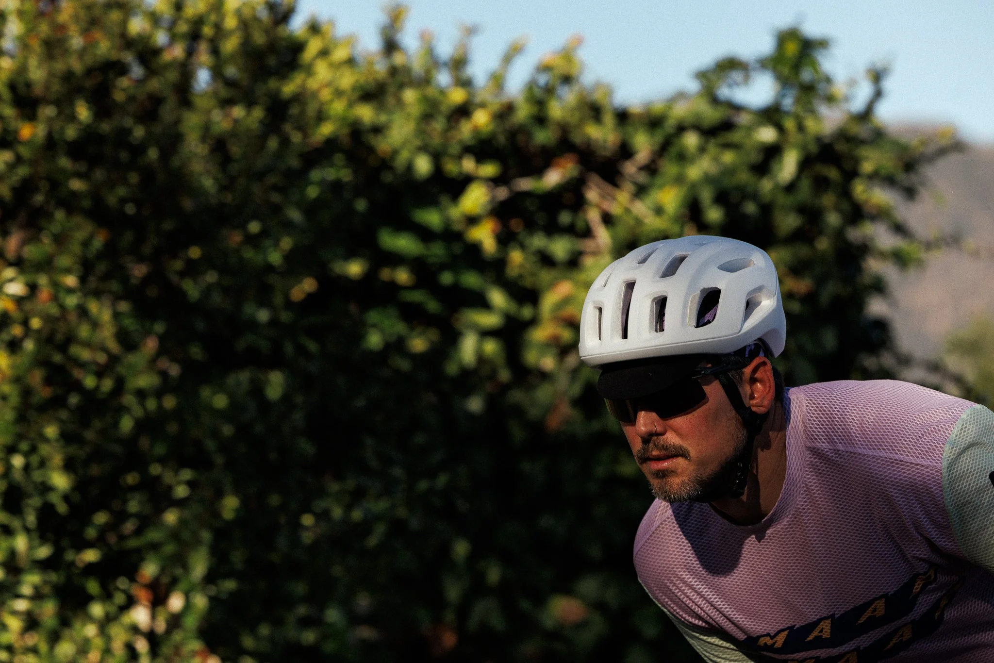 A man wearing a white bike helmet, black sunglasses, and a pink and white cycling jersey rides a bicycle outdoors with green foliage in the background.