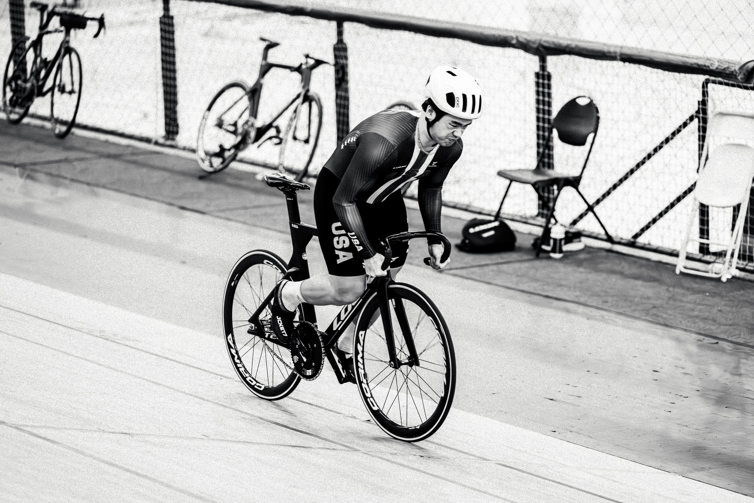 A male cyclist in a USA uniform riding a track bicycle during a race, with bicycles and chairs in the background.