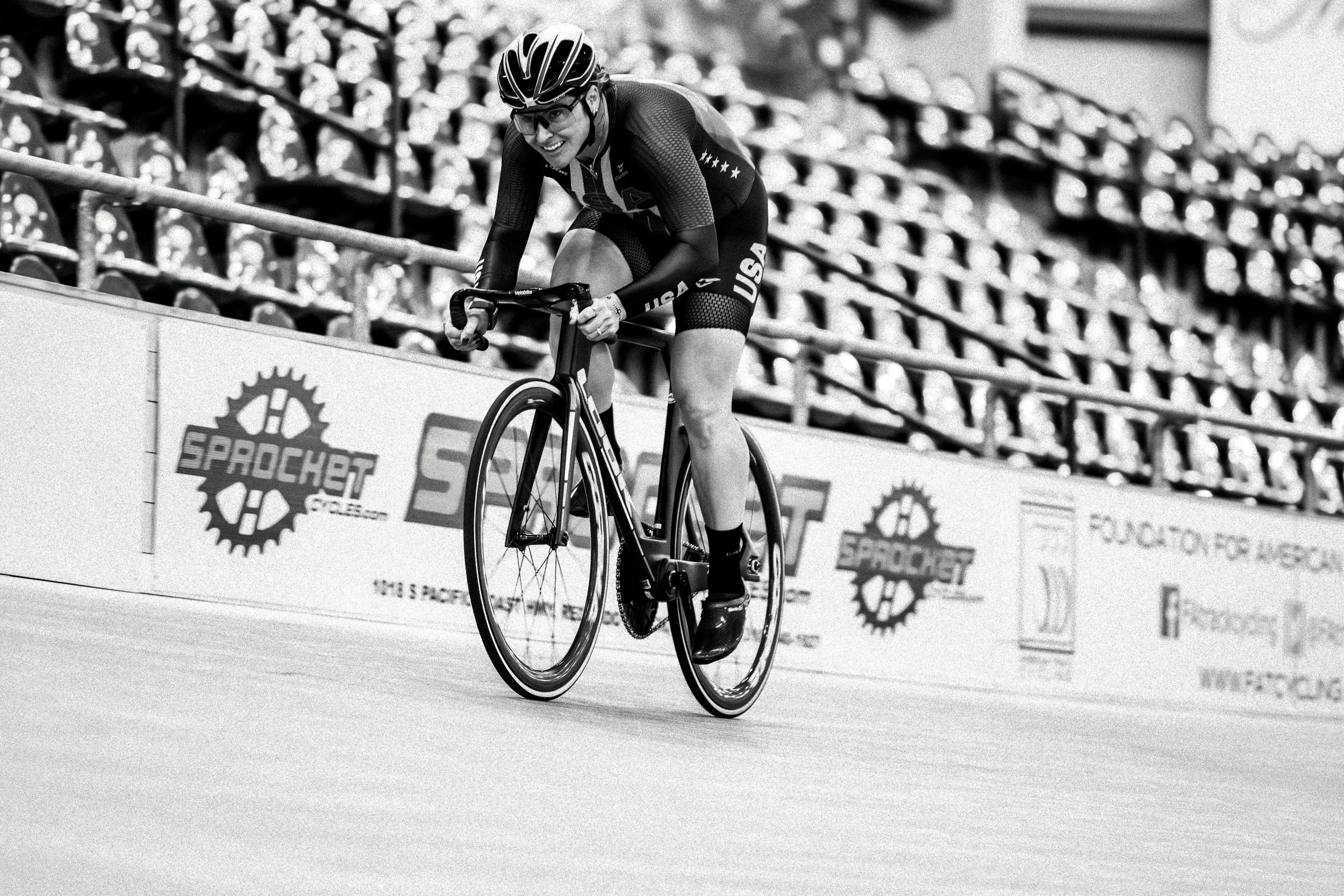 A female cyclist in a USA team uniform, wearing a helmet and glasses, is racing on a velodrome bike track, smiling as she pedals uphill.
