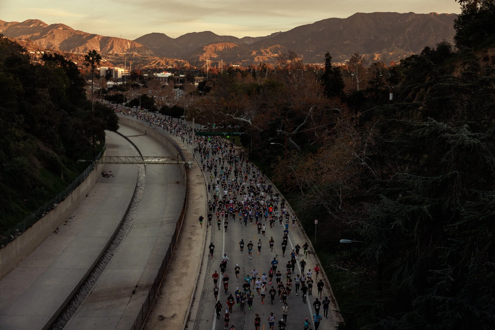 A large group of people participating in a marathon or running event on a road with mountains in the background, during sunset.