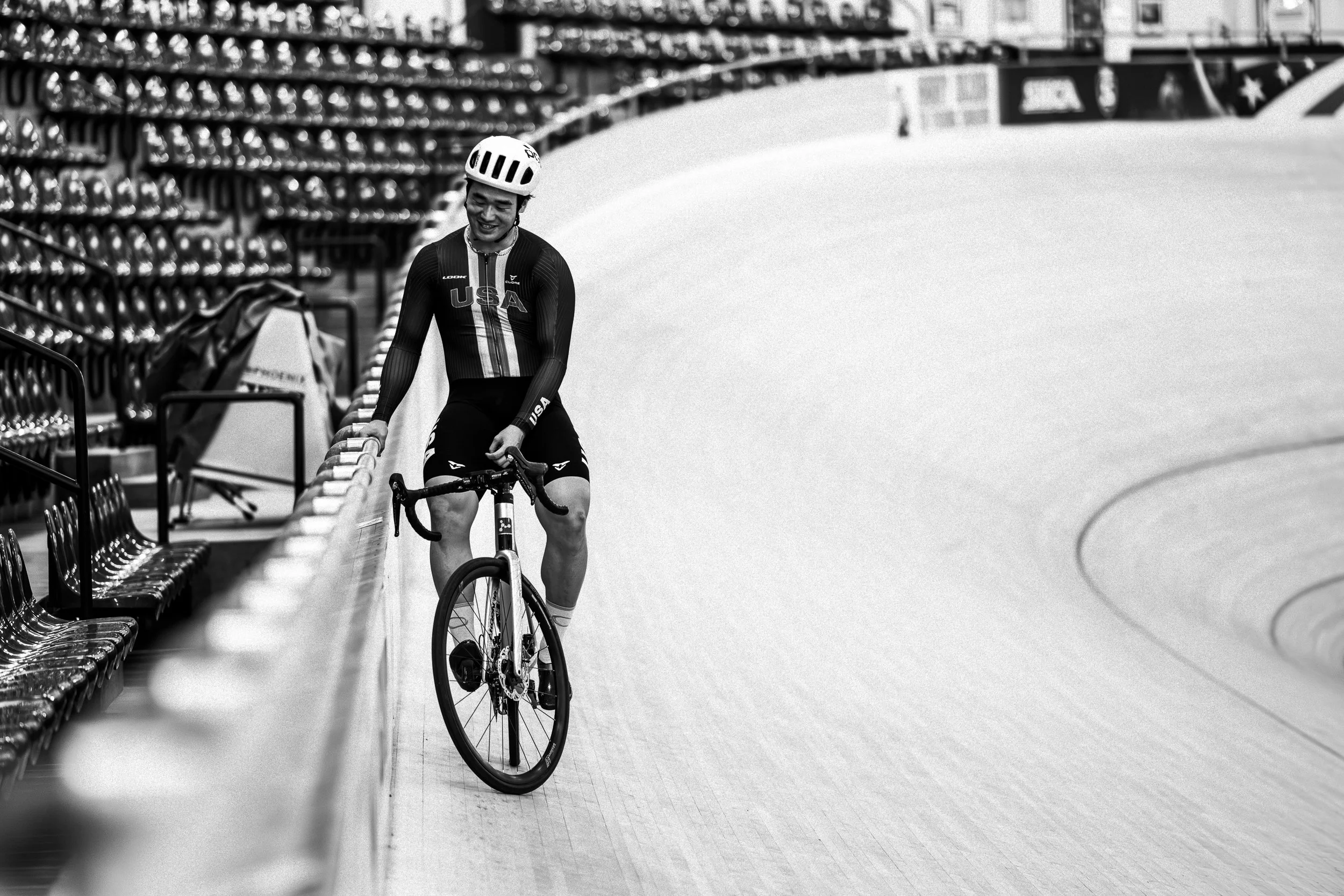 A cyclist wearing a USA team uniform and helmet, sitting on a bike and smiling on an indoor velodrome track.