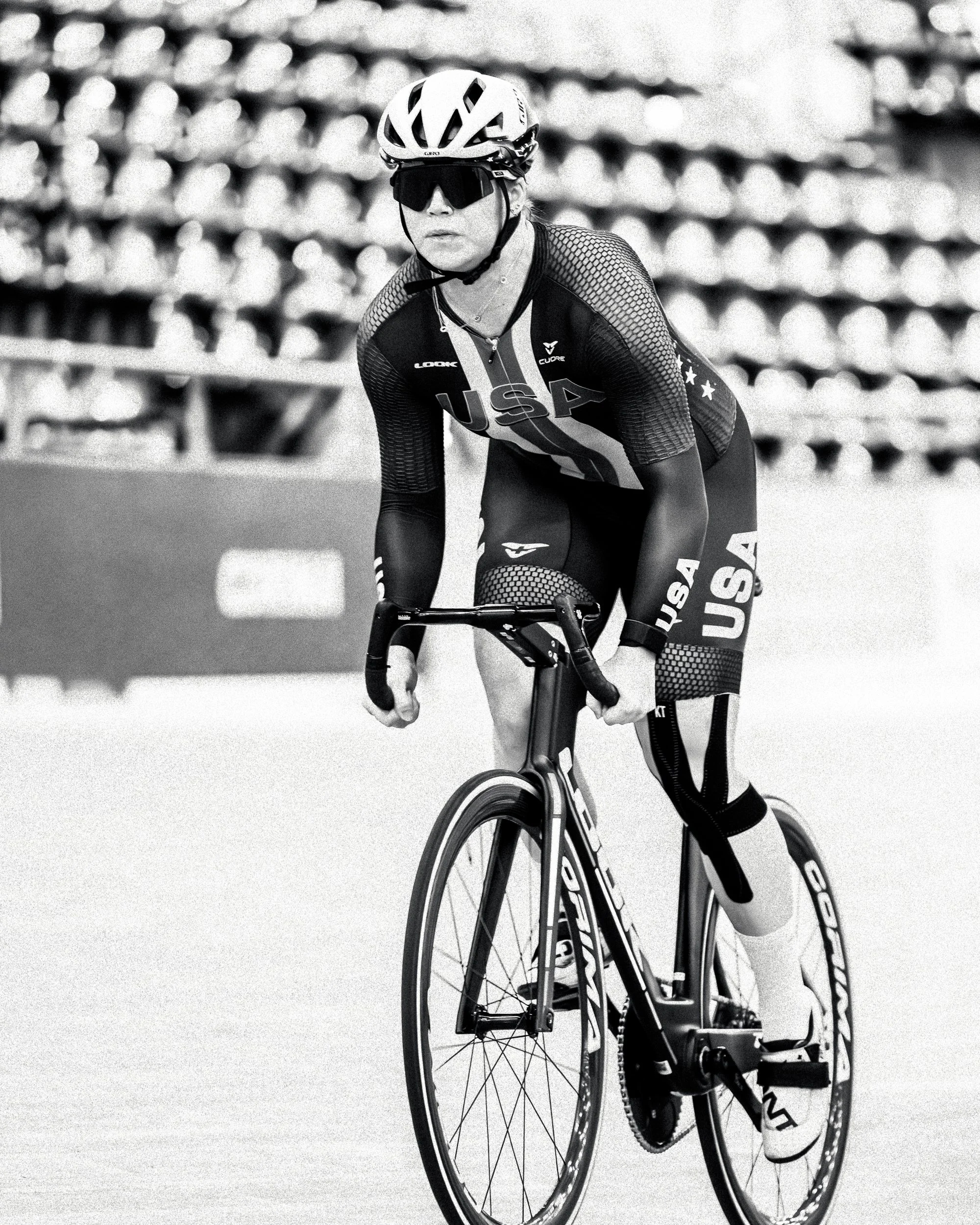 A female cyclist wearing USA team gear, helmet, and sunglasses riding a race bike in a velodrome.