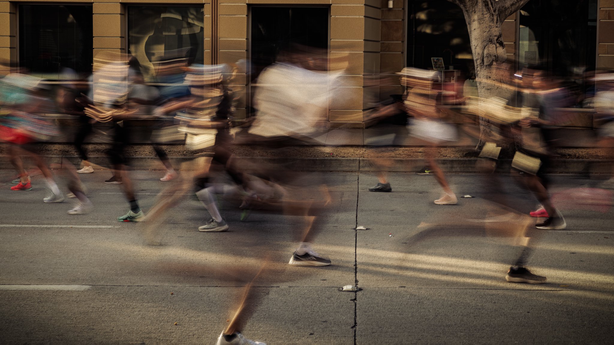 Blurry image of marathon runners running on a city street in front of buildings and a tree.