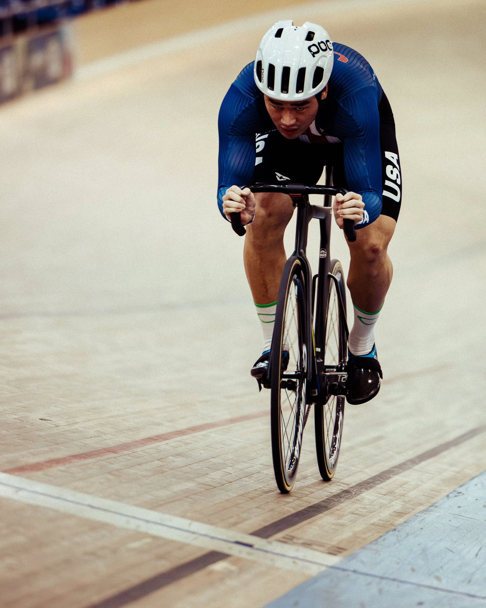 A male cyclist wearing a white helmet and blue USA team uniform, competing on an indoor velodrome track.