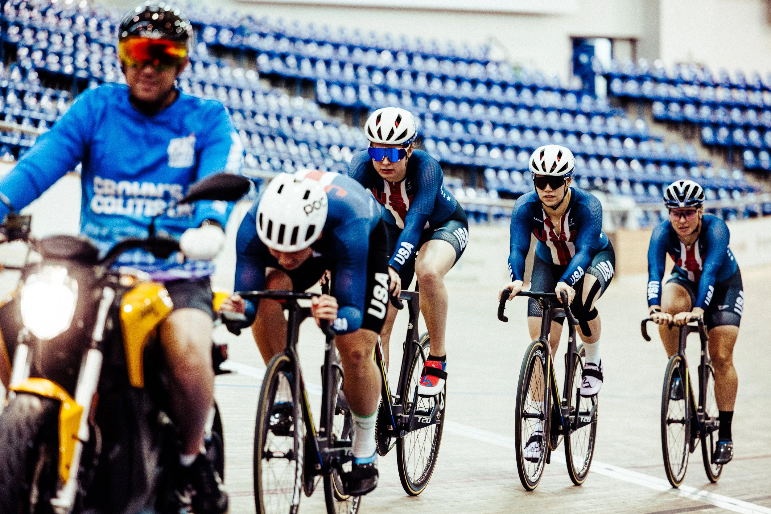 Four female cyclists in USA team uniforms racing on indoor velodrome track with a person on a motorcycle nearby.