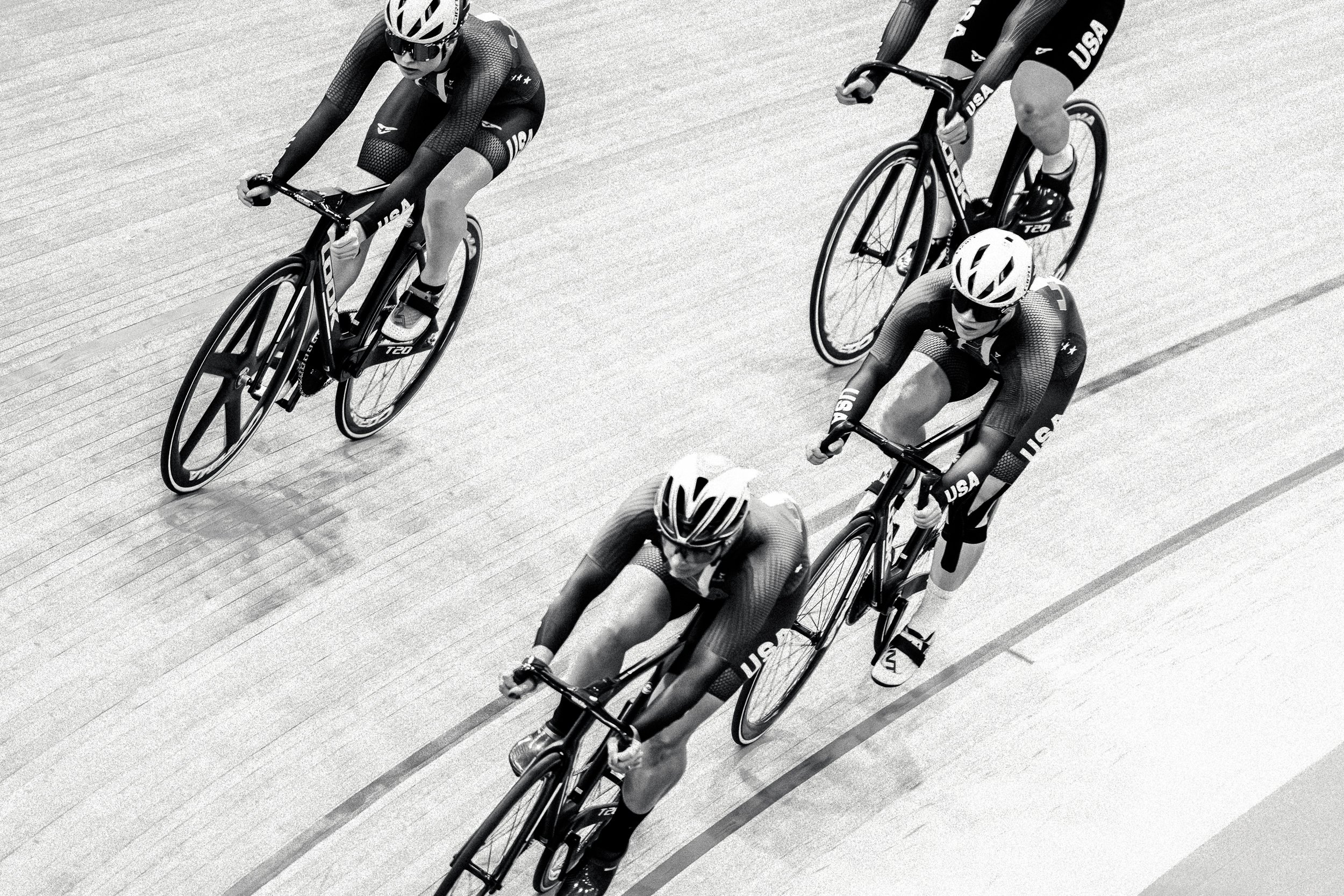 Four cyclists racing on an indoor velodrome track, wearing helmets and USA team uniforms.