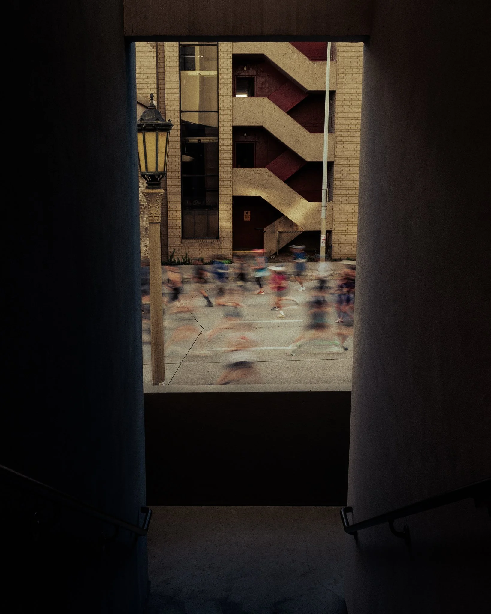 Through a window, a blurred crowd of runners participating in a race on a city street, with a brick building, stairwell, and street lamp visible in the background.