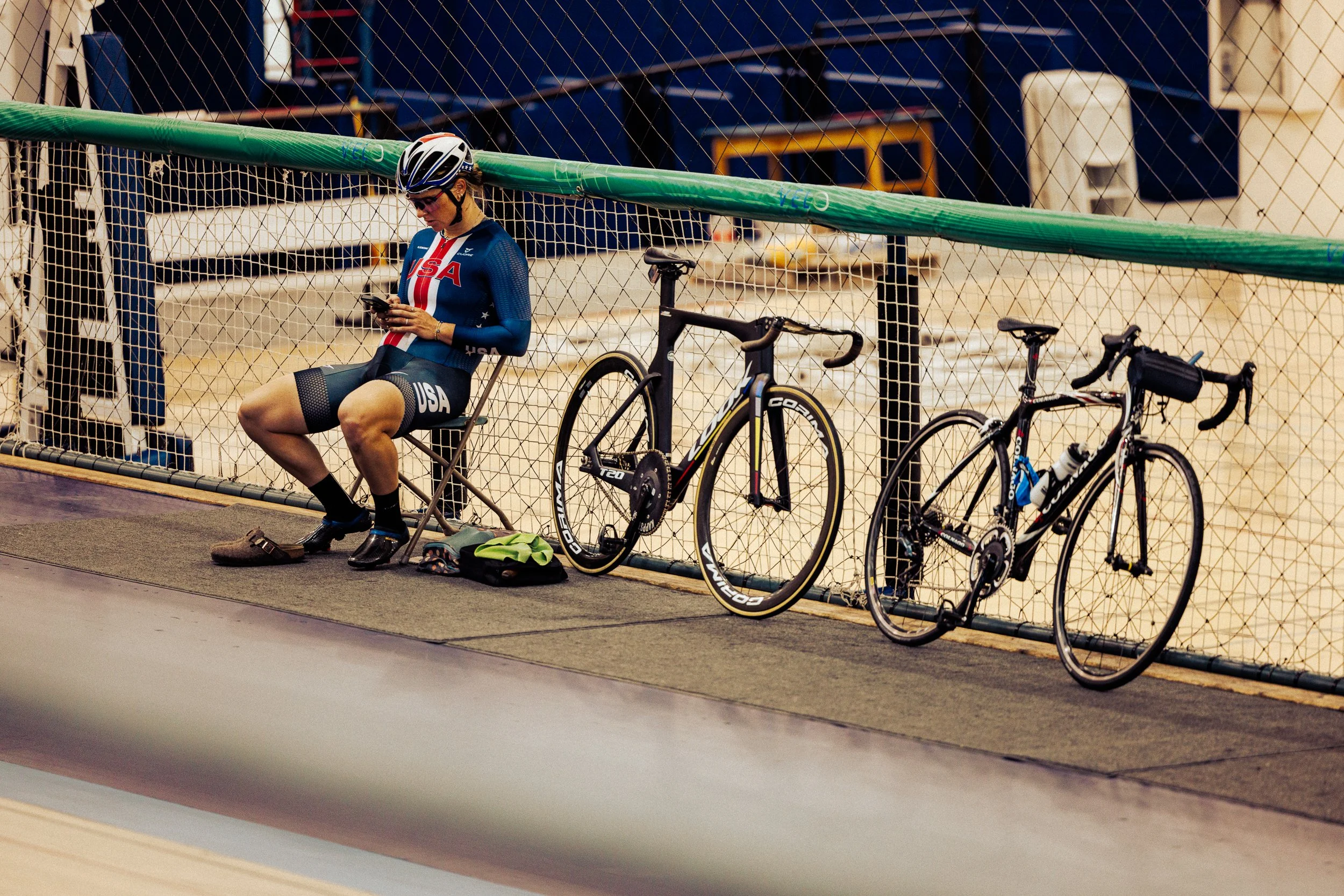 A female cyclist in USA team gear sitting on a folding chair indoors, looking at her phone, with two racing bicycles leaning against a chain-link fence beside her.