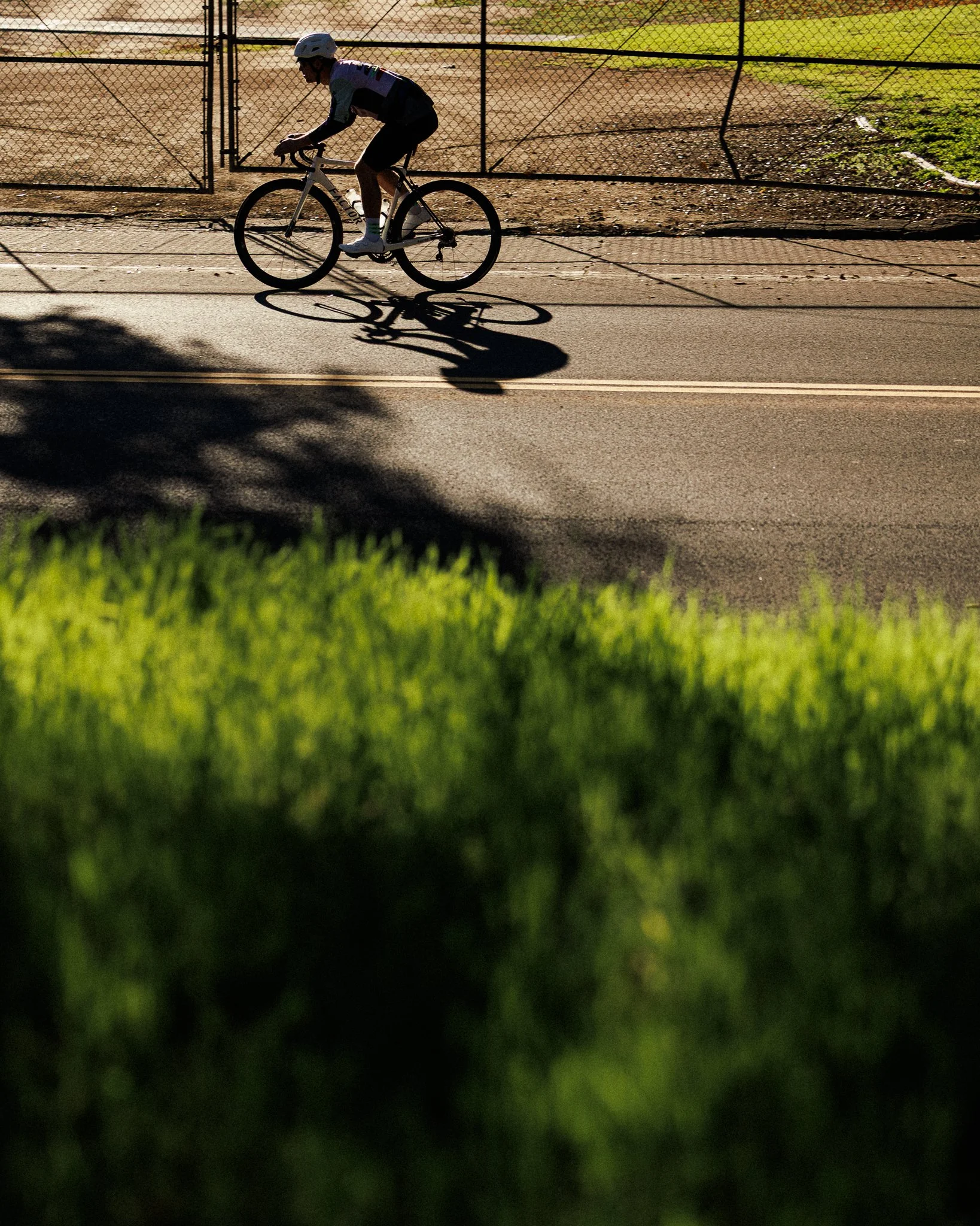 A cyclist wearing a helmet riding a bike on a paved road, with the shadow of the cyclist cast on the ground, near a fenced area and green grass in the foreground.