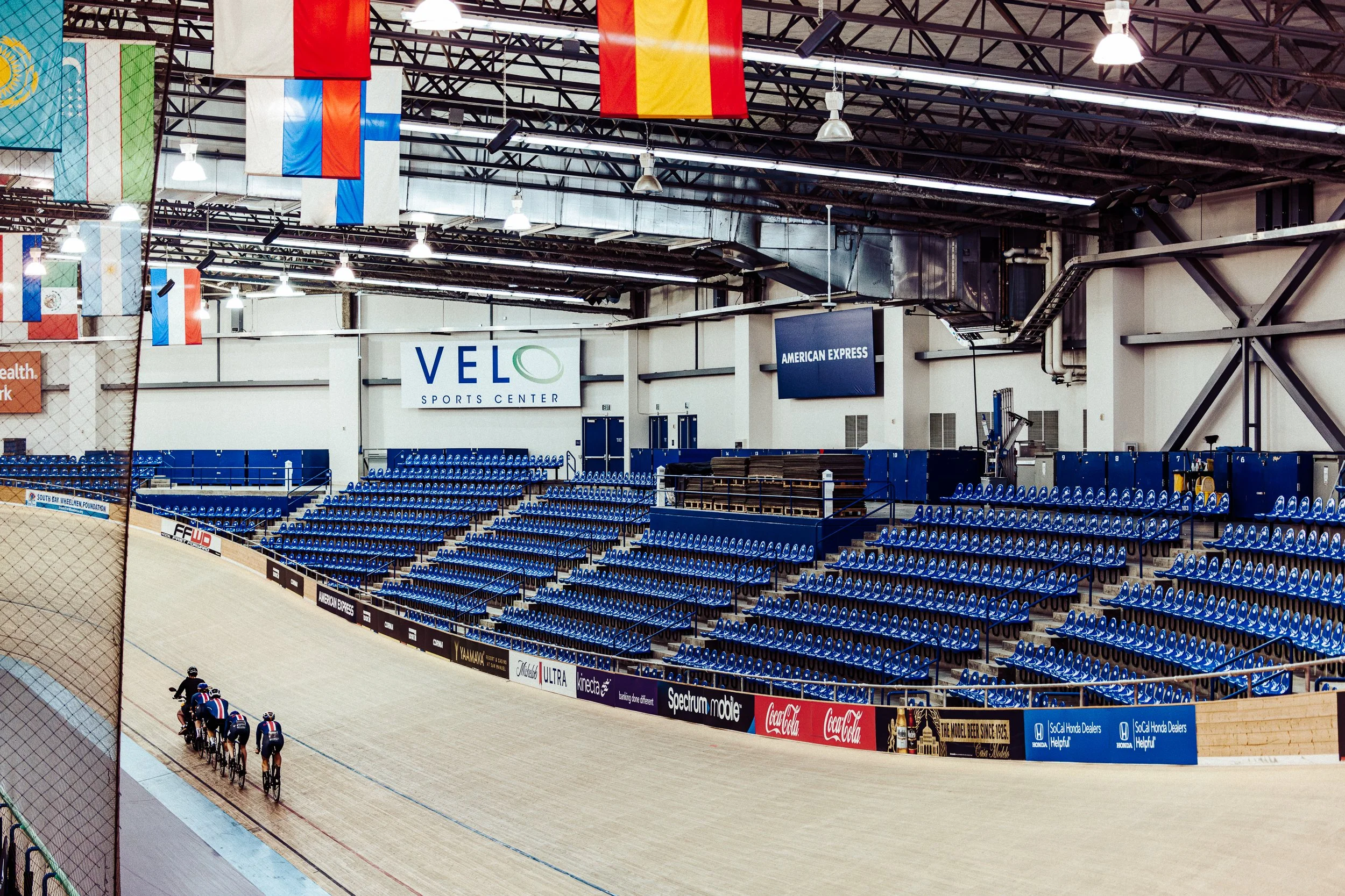 An indoor velodrome track with a team of cyclists riding together. The stadium has blue seating and banners hanging from the ceiling, including flags from different countries and an American Express sign. The velodrome is part of the VELO Sports Cent