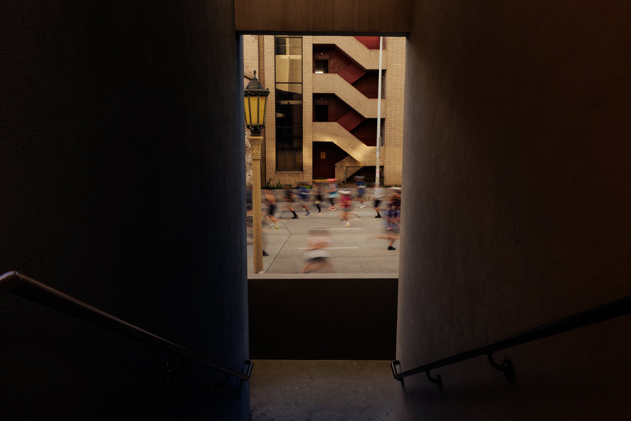 View through a rectangular opening in a dark stairwell showing blurred runners crossing the street, with a building, a streetlamp, and an alley in the background.