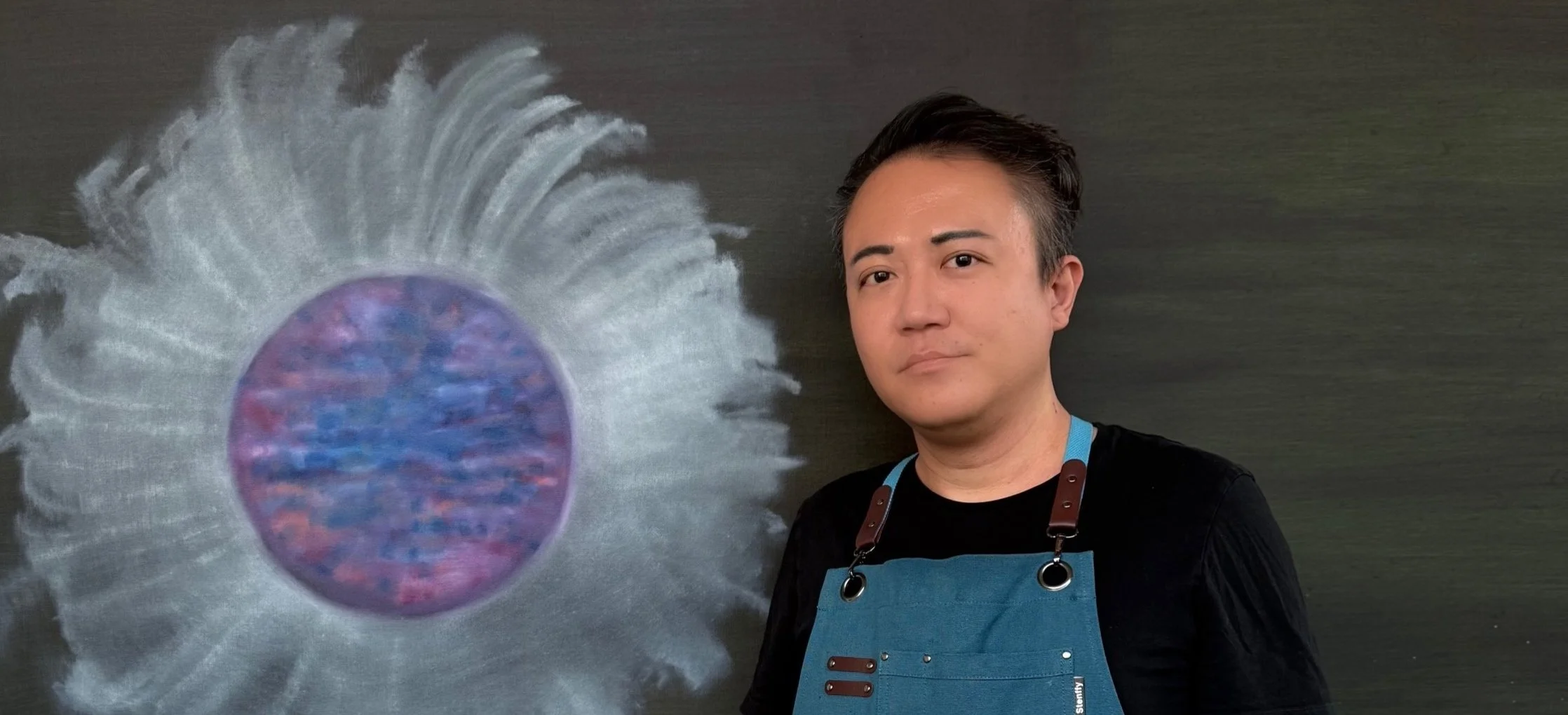A man with short dark hair and a serious expression wearing a black shirt and blue apron standing next to a colorful abstract artwork of a circular shape with white streaks around it on a dark background.