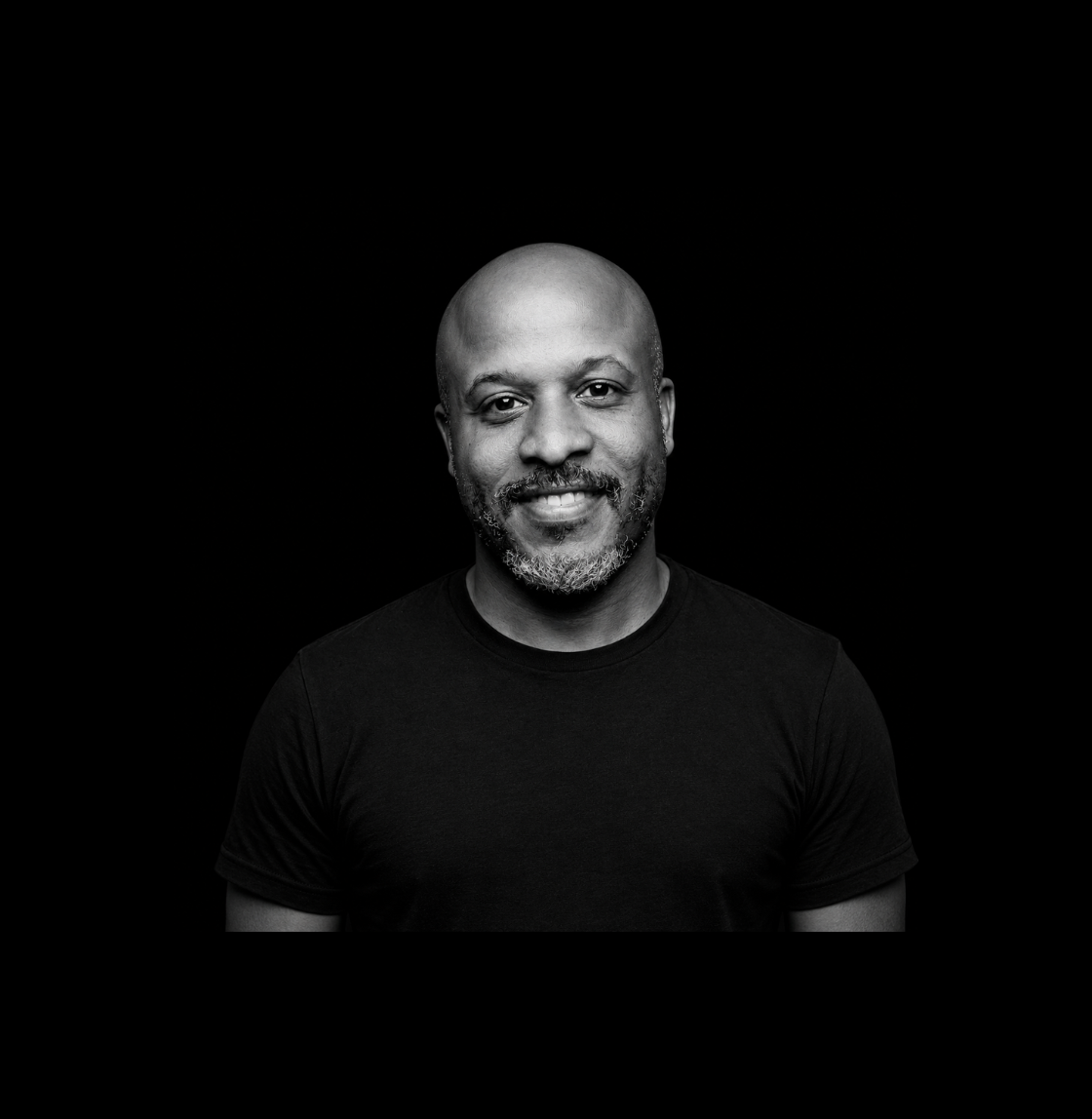 A Black, bald man with graying beard in a black t-shirt against a black background
