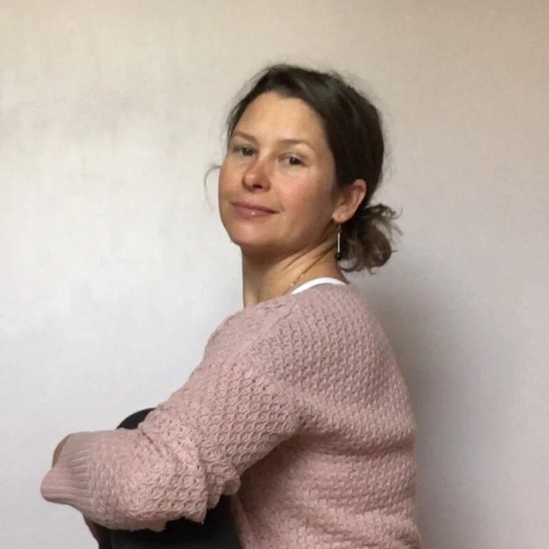 A woman with dark hair tied back, smiling softly, wearing a light pink textured sweater and a white shirt underneath, standing against a plain, light-colored wall.