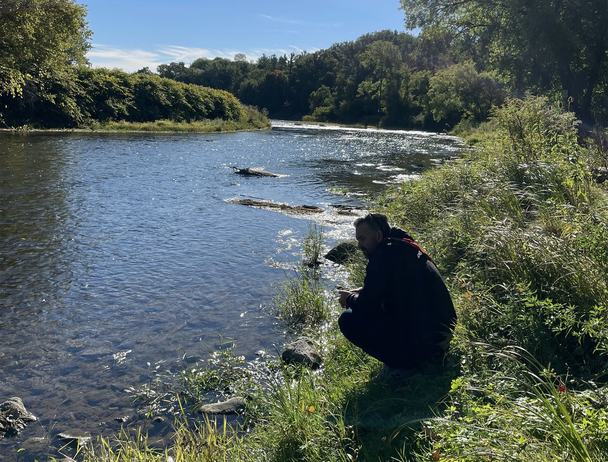 A person sitting near the edge of a river with trees and clear blue sky in the background. The person appears to be looking at their phone.
