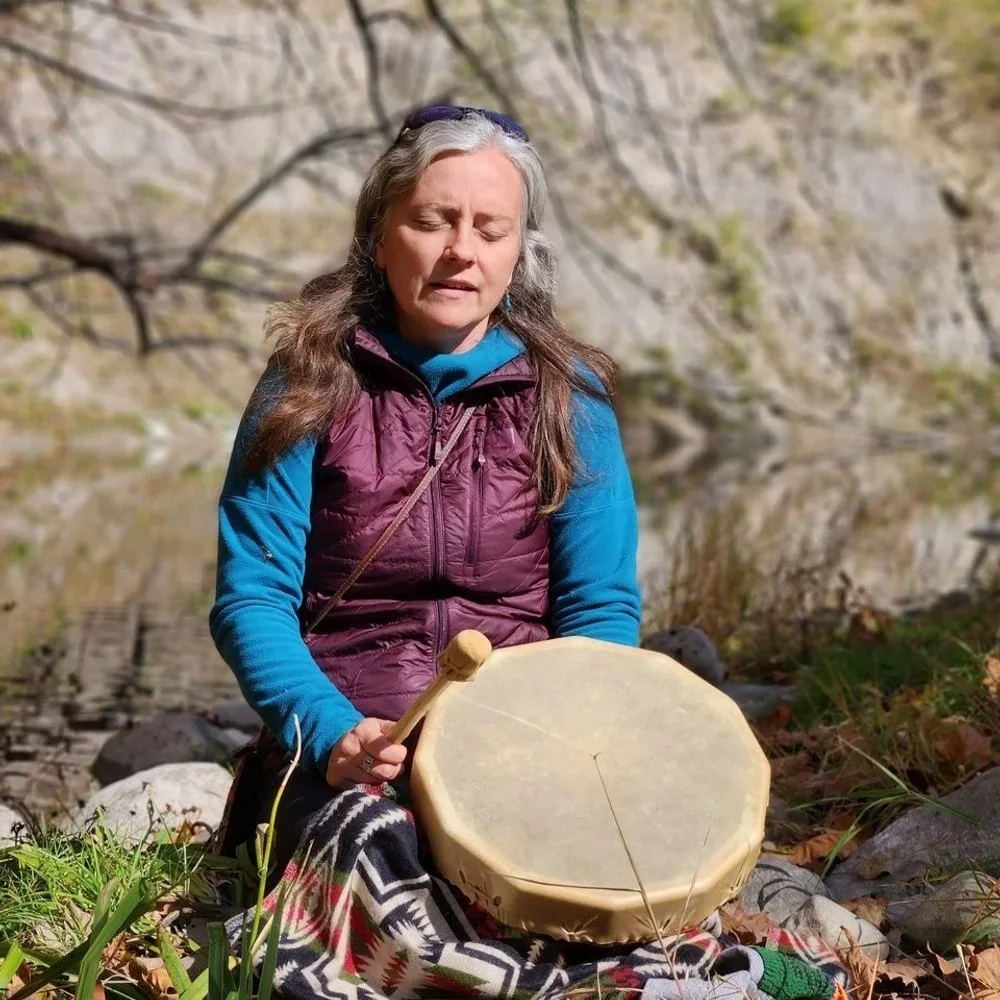 A woman with gray hair, wearing a purple vest and blue long sleeve shirt, is sitting outdoors near a body of water, holding a drum and a mallet, with her eyes closed.