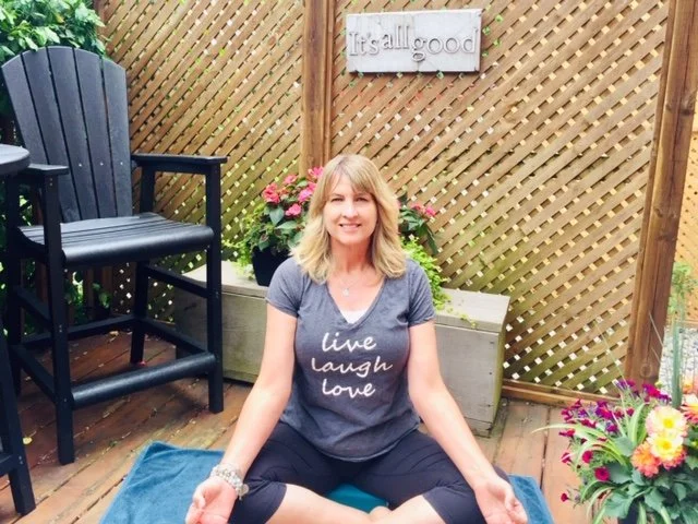 A woman is seated on a blue mat in an outdoor wooden deck area, smiling at the camera. She is wearing a gray t-shirt with the words 'live laugh love' and black pants. Behind her, there are potted flowers and a decorative sign that reads 'It's all good' on a lattice fence.