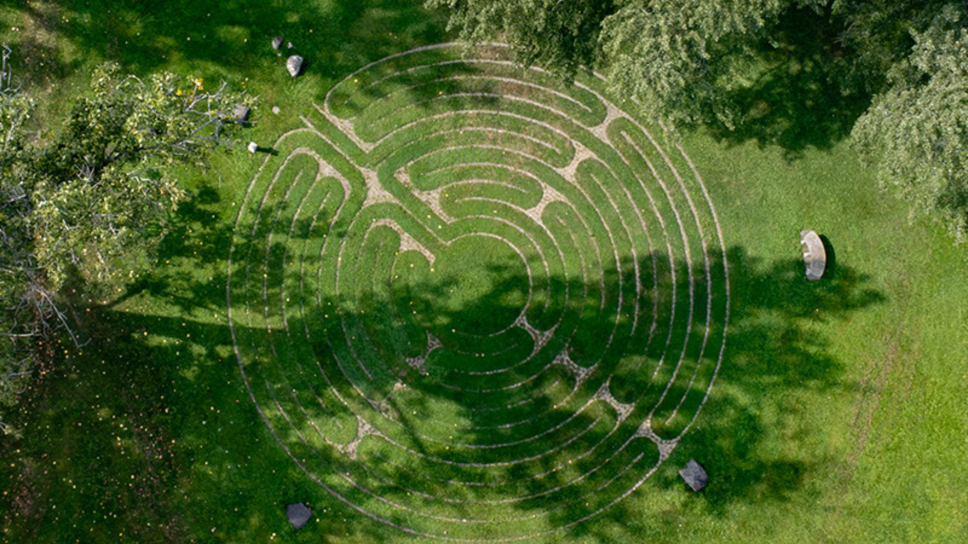 Aerial view of a labyrinth maze with curved paths made of gravel or stones in a grassy park with trees and a small white circular structure nearby.