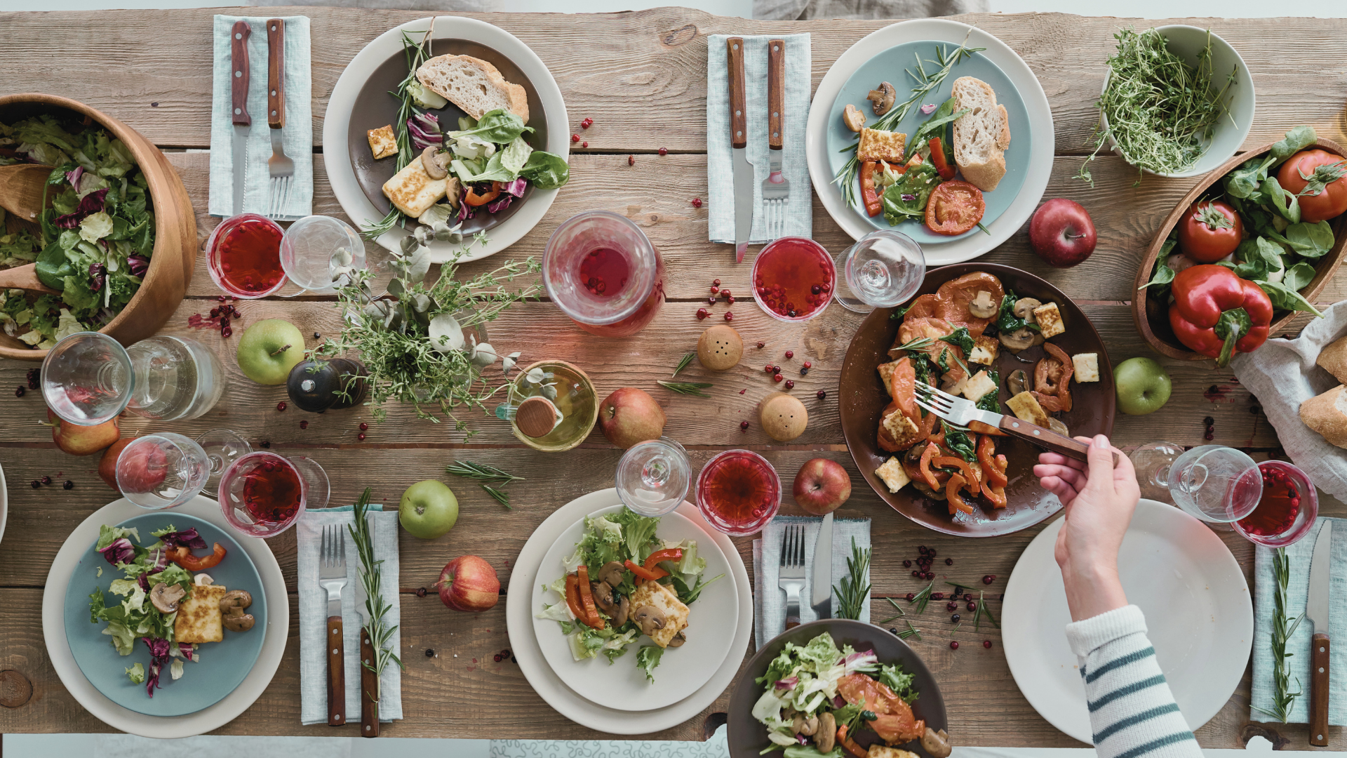 A top-down view of a wooden dining table set for a meal with salads, bread, grilled vegetables, apples, tomatoes, and glasses of red wine, with a hand reaching to serve food from a plate.