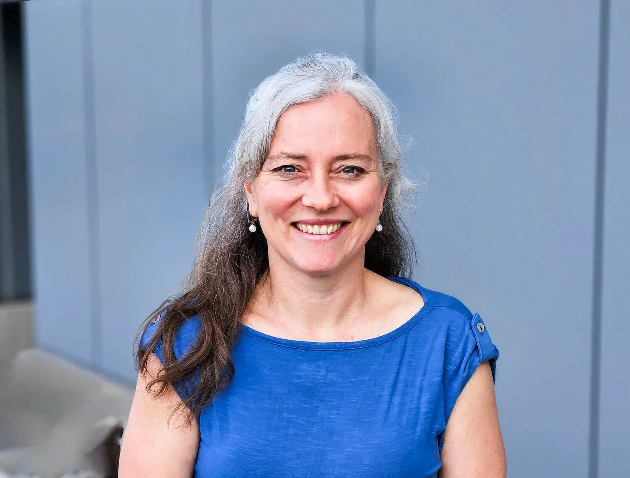 A smiling woman with gray hair, wearing a blue top and pearl earrings, standing outdoors against a gray wall.