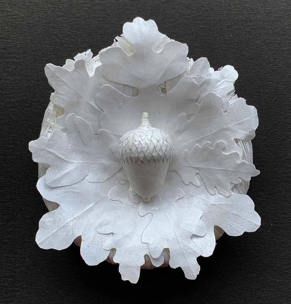 handmade 3D paper sculpture titeld "Acorn Oak" featuring a paper acorn cradled in hand-cut white oak leaves mounted on a wood base, ready to hang.