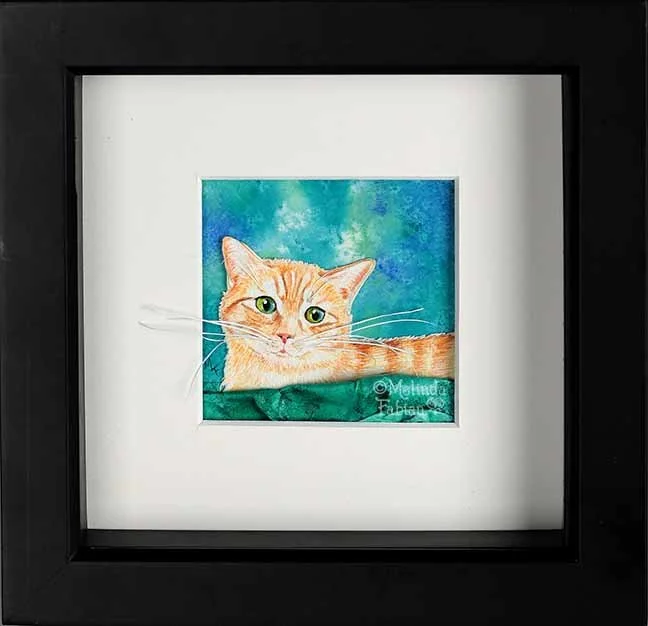 paper scupture cat - and orange ginger tabby cat is sitting on a 3D paper sculpted teal colored blanket. The hand painted watercolor cat has bright green eyes. The cat has long 3D whiskers that extend over the white mat of the picture.
