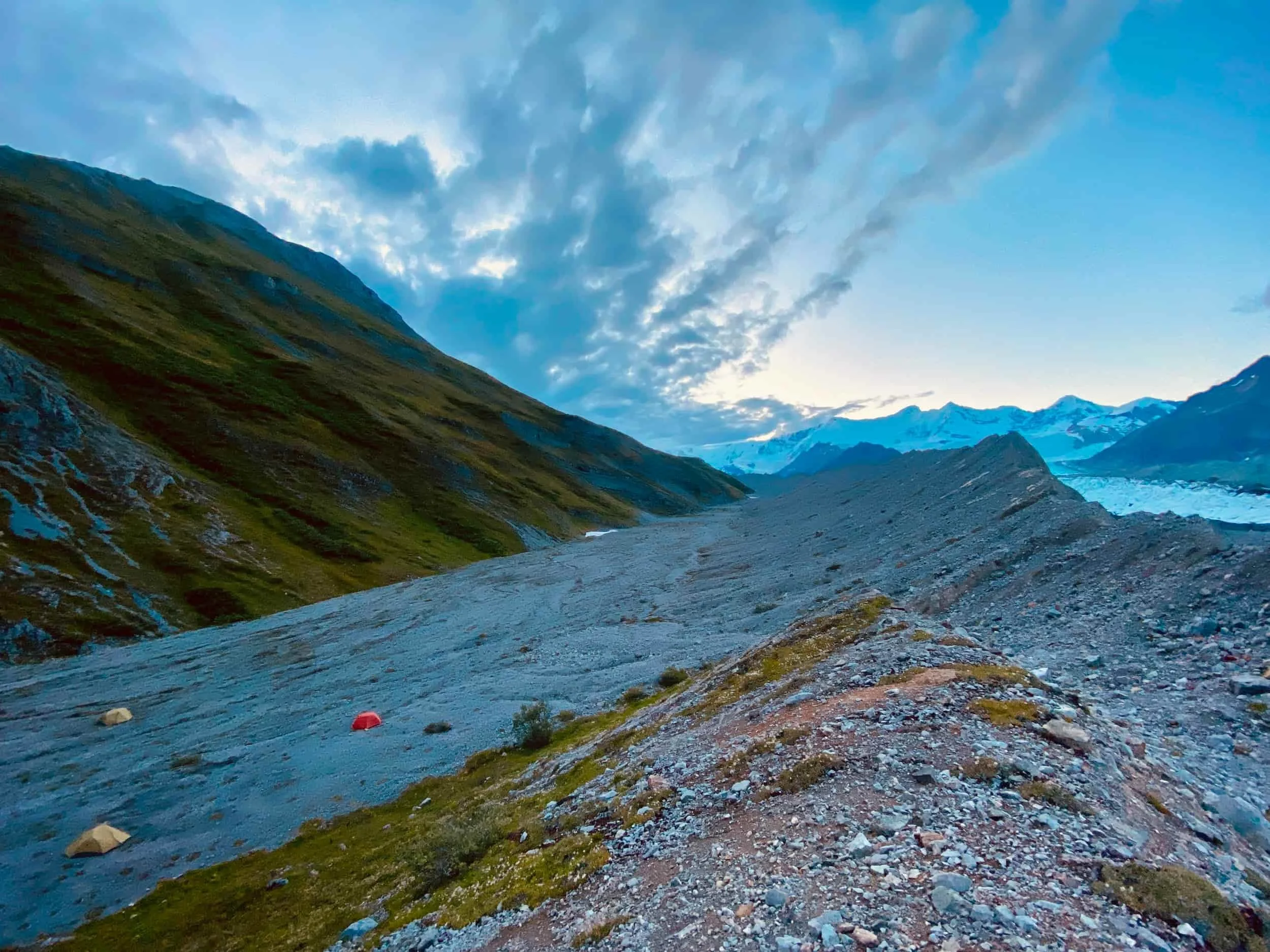 Tents in a gravel valley in Alaska with snowcapped Wrangell Mountains behind.