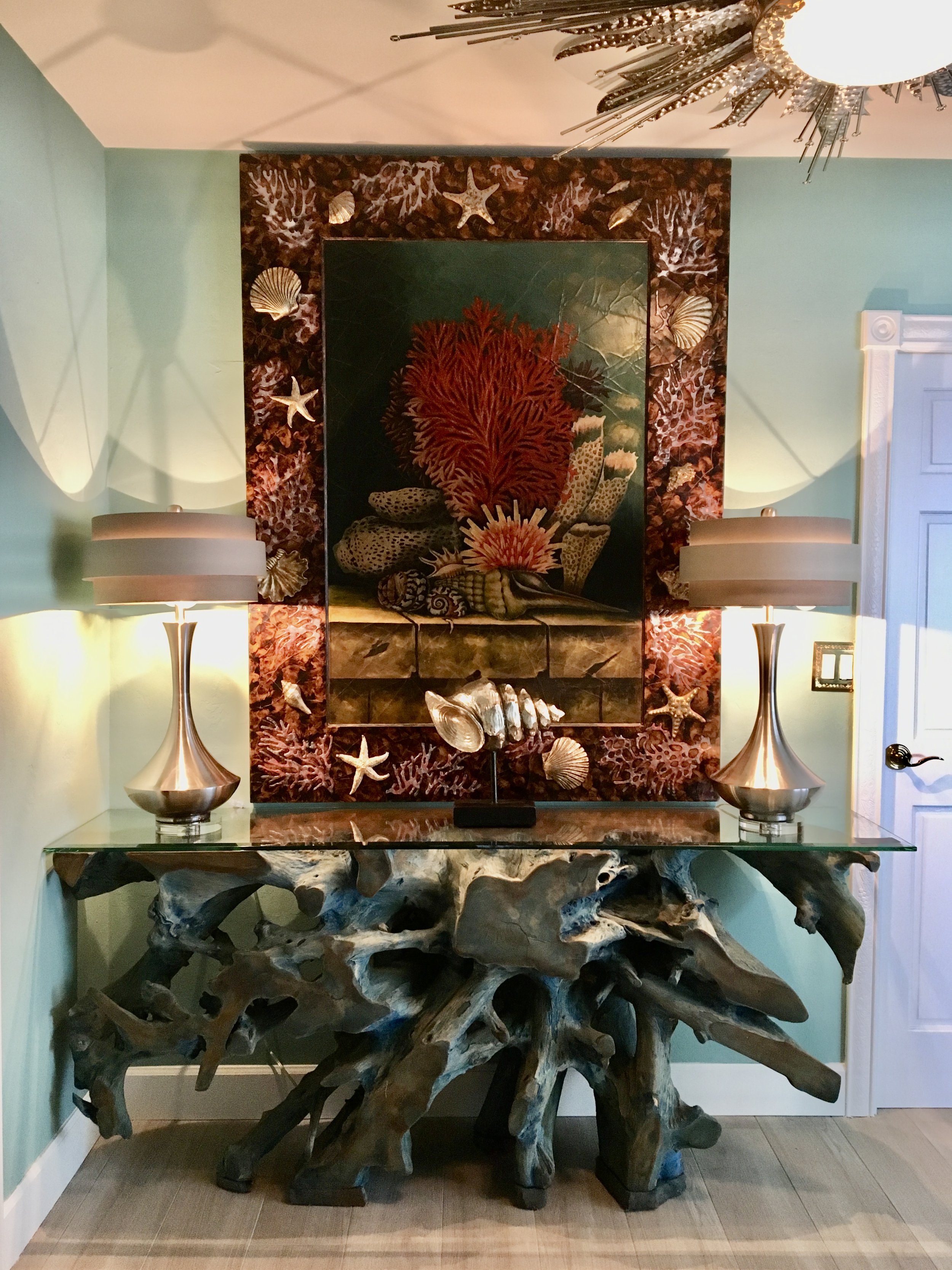 Luxury coastal interior design featuring a sculptural driftwood console table with glass top, coral artwork, seashell accents, and designer table lamps in a Fort Lauderdale home
