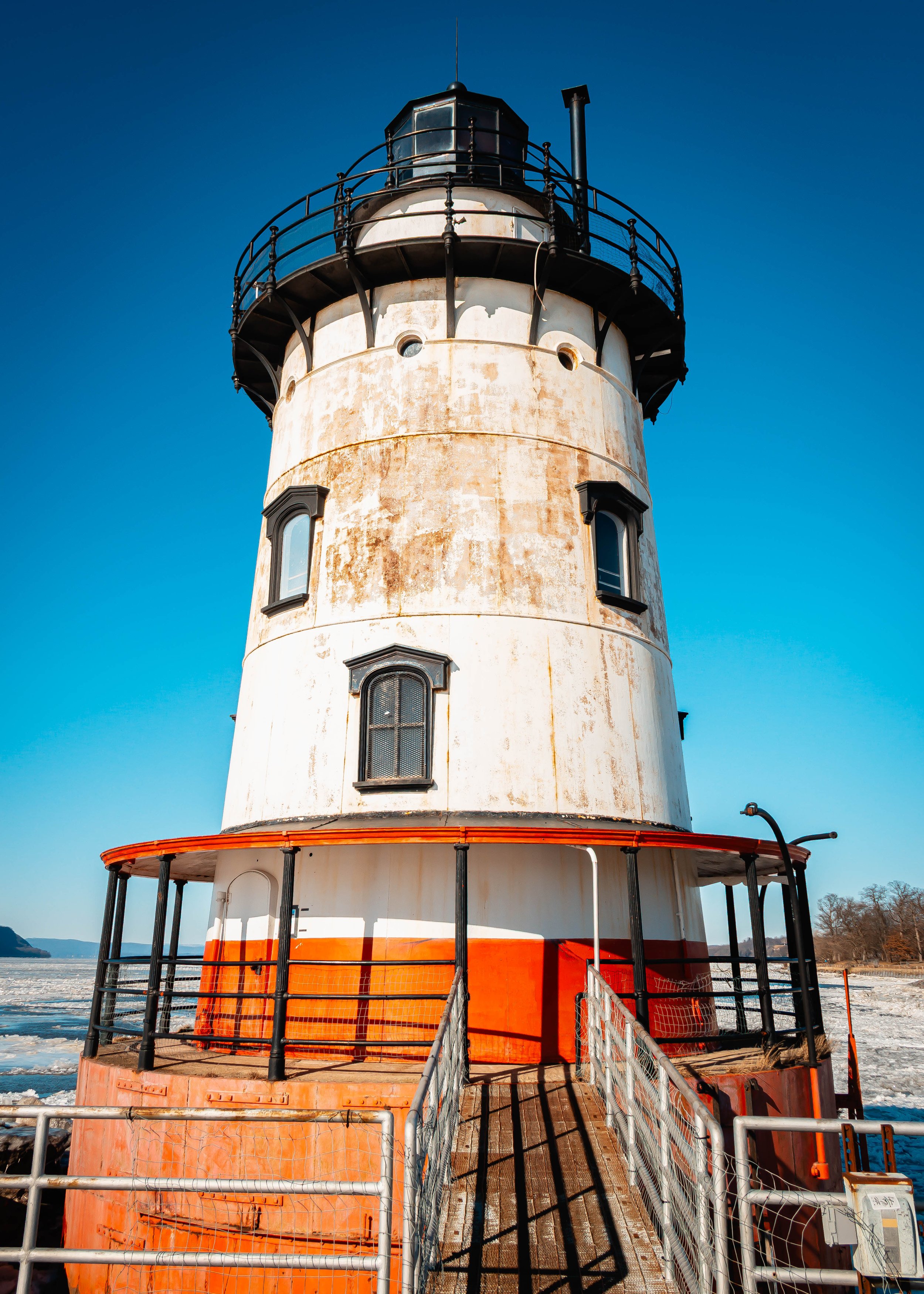 A tall lighthouse with a weathered white and orange exterior, black railing around the top, and a small dock at the base, against a clear blue sky.