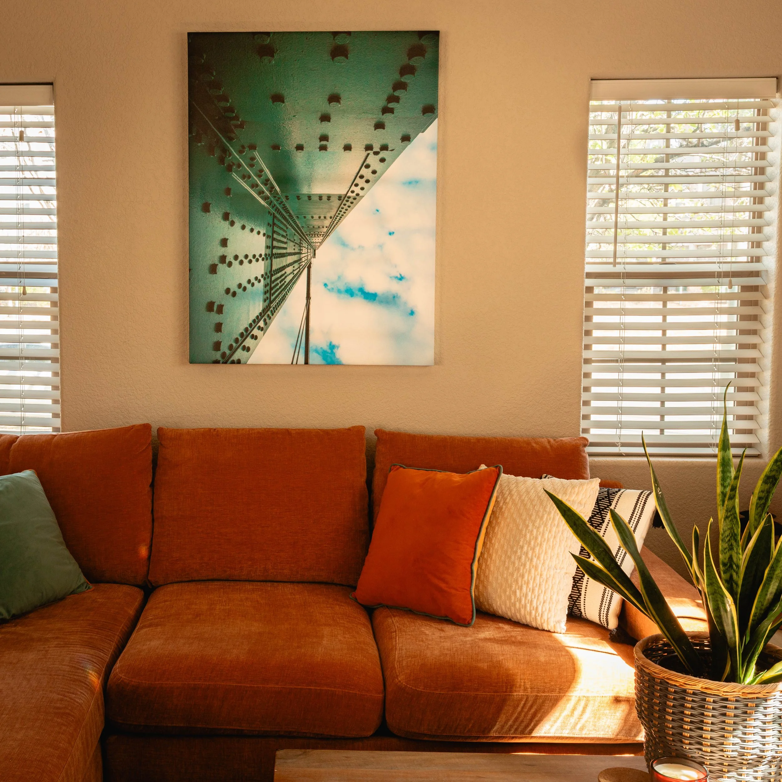 A living room with a brown velvet sofa adorned with orange and cream pillows, a large artwork of a tower viewed from below, three windows with white horizontal blinds, and a plant in a woven basket on a small table with a candle.
