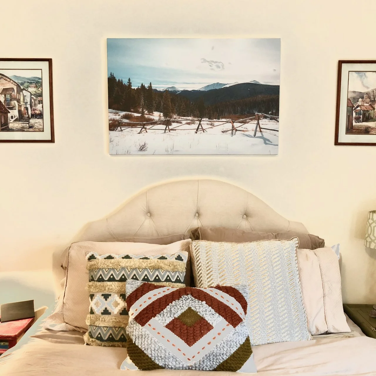 A cozy bedroom with a beige tufted headboard, decorated with patterned throw pillows, and framed landscape paintings hanging on a cream-colored wall.