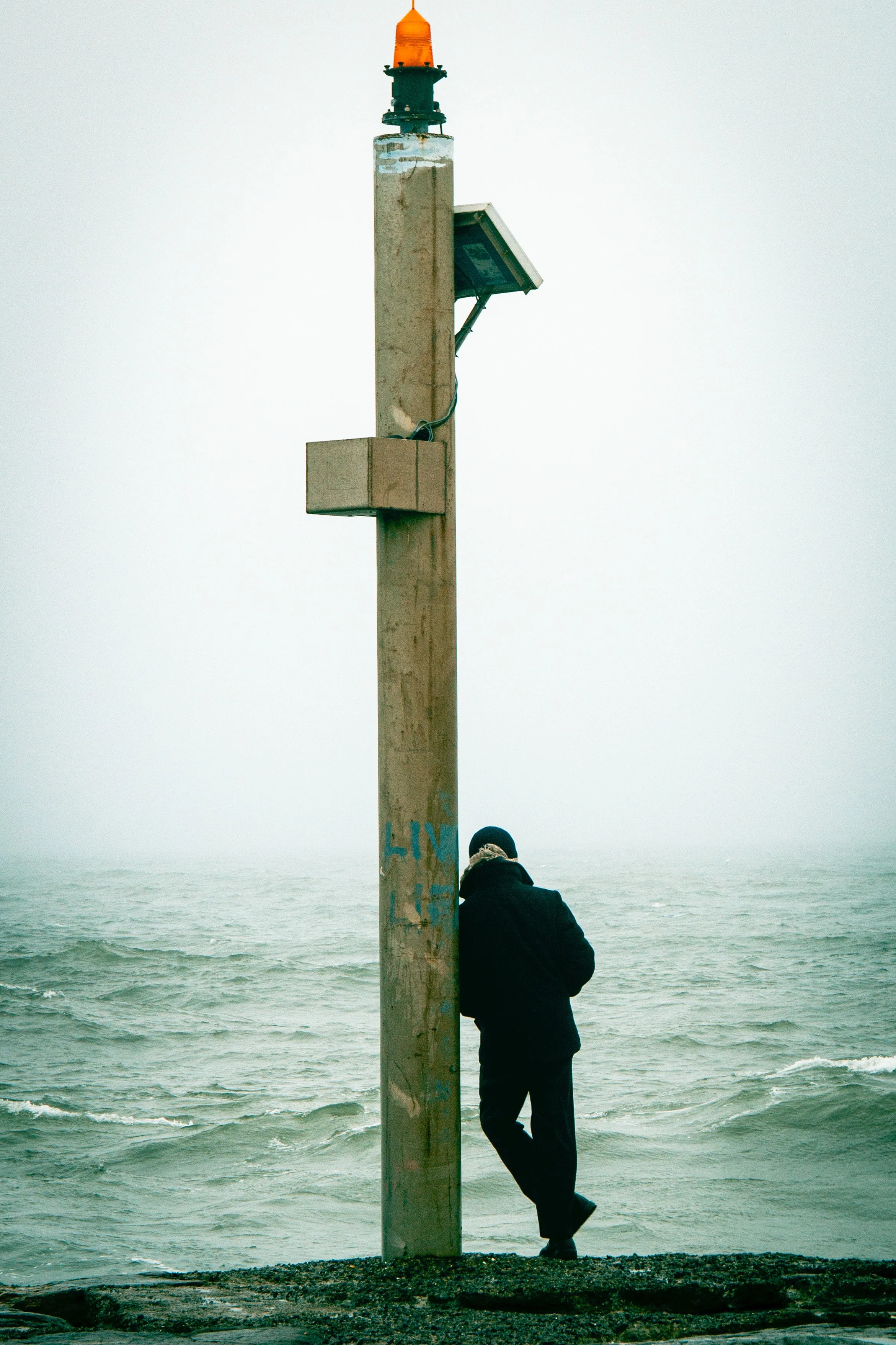 A person leaning against a concrete utility pole on a beach, with the ocean and foggy sky in the background. The pole has graffiti and a solar panel attached to it.