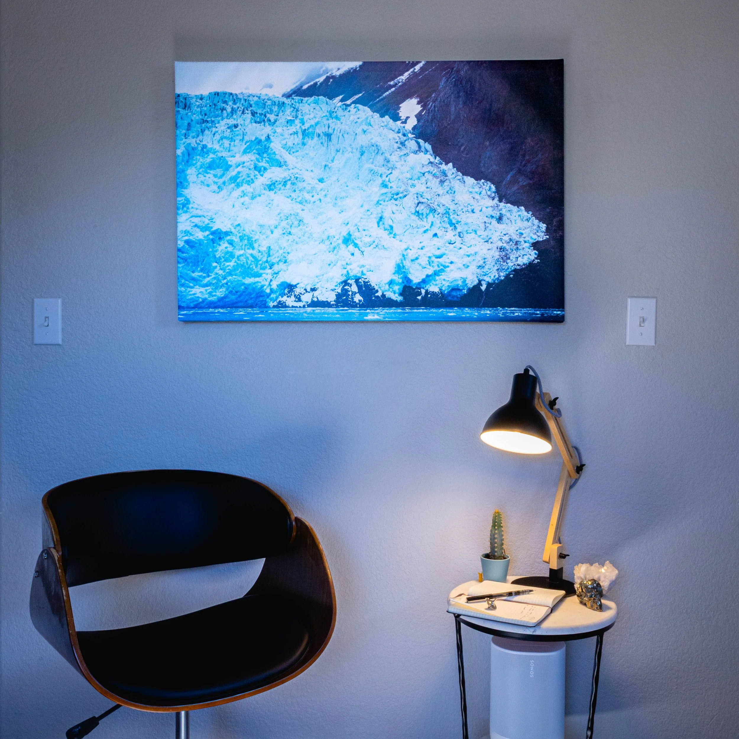 A wall with a framed mountain and glacier photograph, a side table with a potted cactus, a notebook with a pen, a decorative object, a desk lamp, and two electrical outlets.