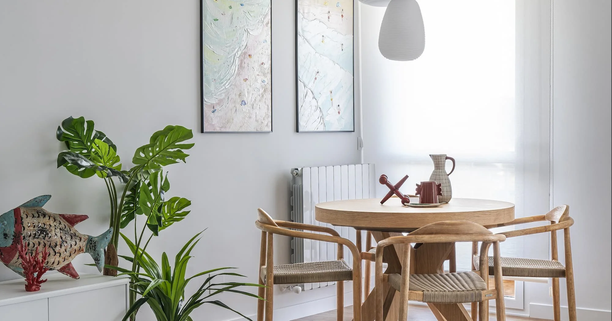 Luminosa zona de comedor junto a ventana con mesa de diseño de madera redonda y sillas de madera con asiento de cuerda en tonos neutros, lámparas colgantes blancas y en la pared dos cuadros  con motivos veraniegos  de bañistas en tonos pastel.