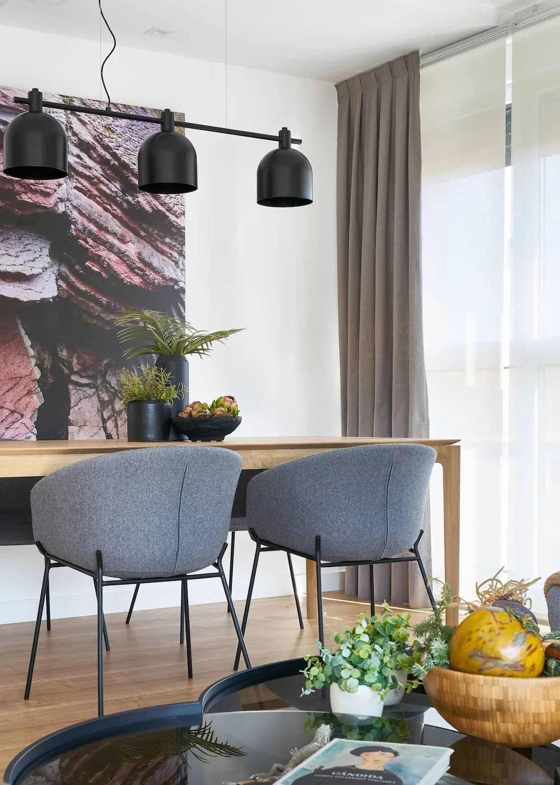 Proyecto de interiorismo en Urdúliz. Salón comedor donde destacan las sillas tapizadas en tela gris y la mesa de madera de Roble natural. Para la iluminación lámpara colgante negra contrasta con una fotografia de gran formato que muestra texturas y c