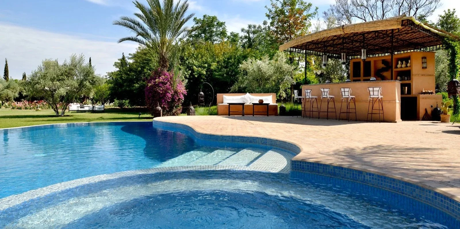 Poolside area at a Yogalates London retreat, with a pool, cushioned lounge area and an outdoor bar surrounded by trees and greenery.