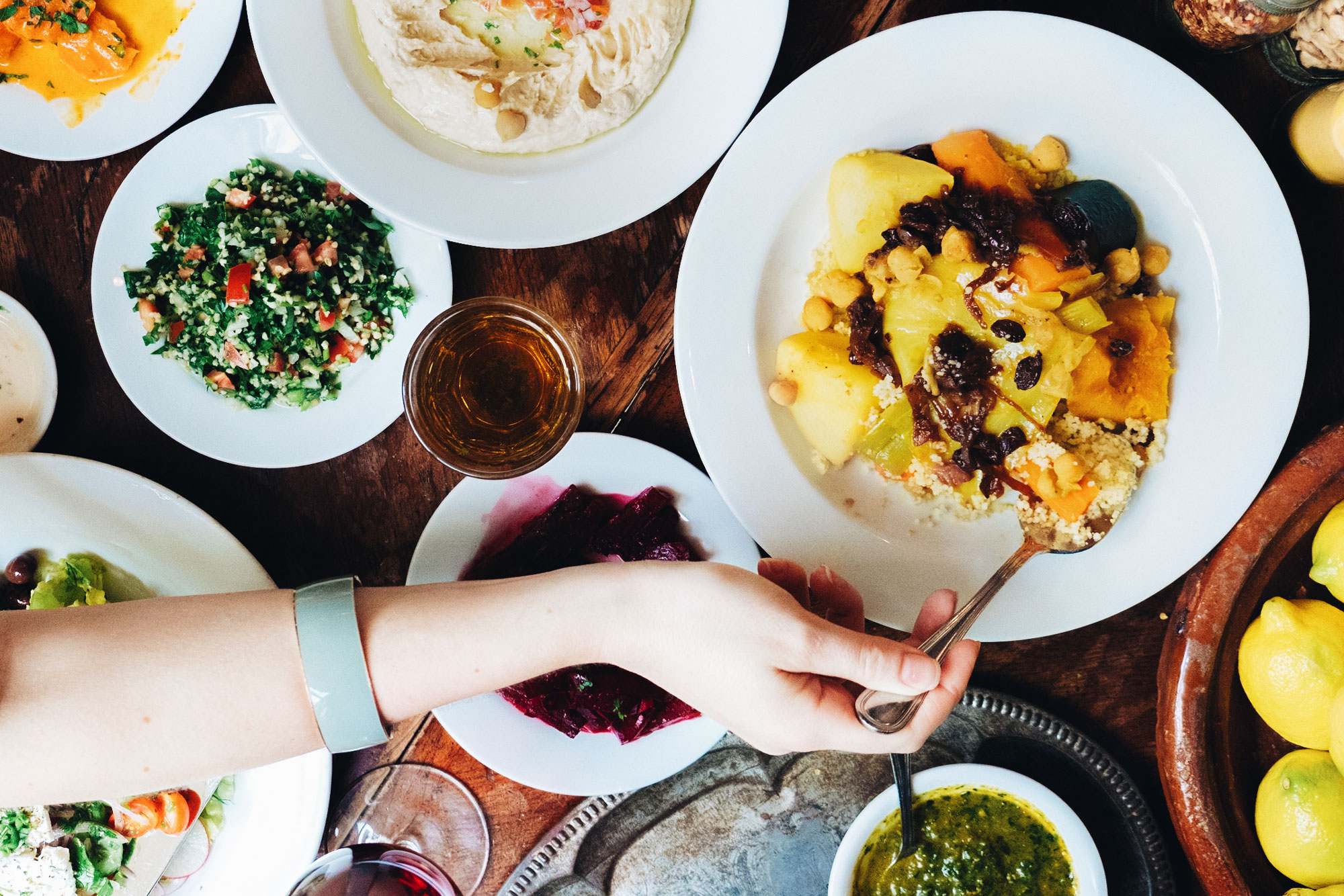 A womans hand reaching for the vegetable couscous. Hummus platter, tabouli, beets and lemons on the table.